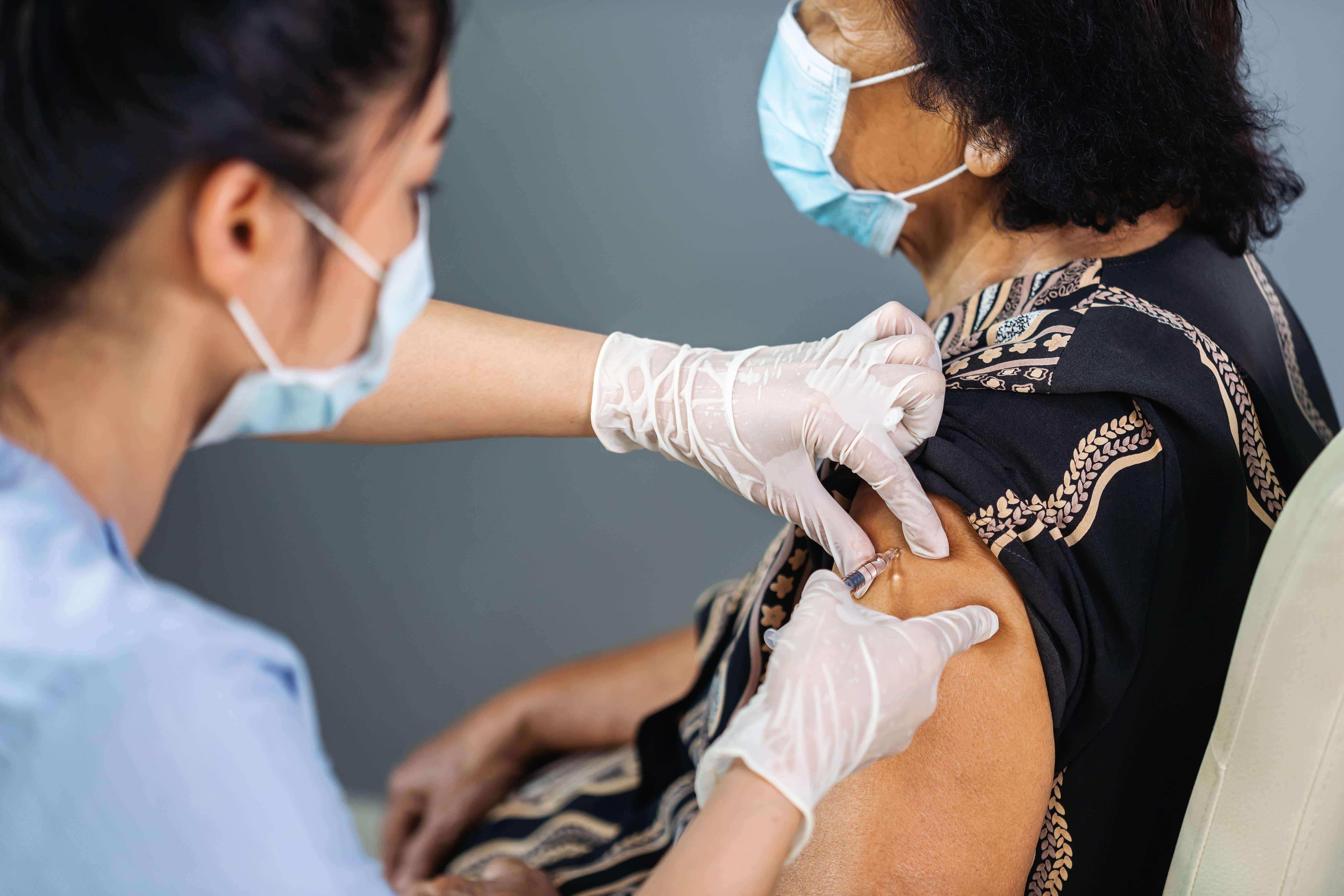 Healthcare worker wearing gloves administering vaccine to an older woman wearing a face mask.