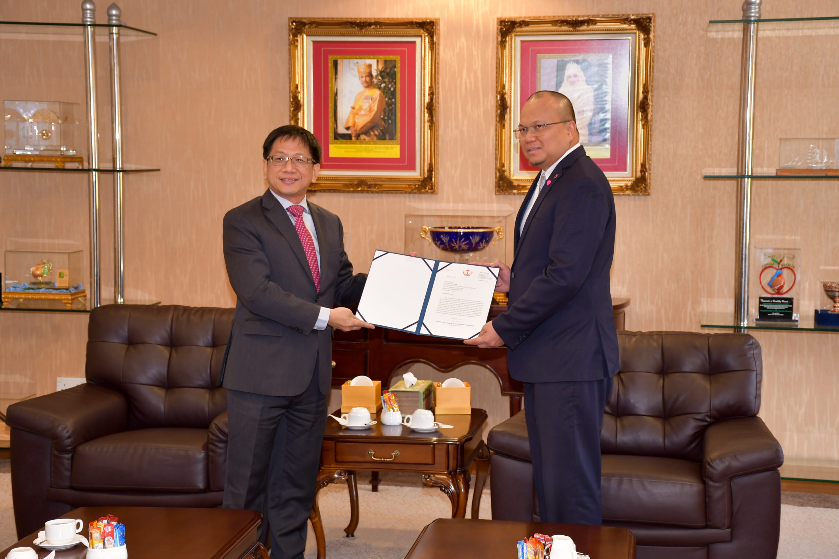 Two men in suits holding a document in an office with portraits and shelves.