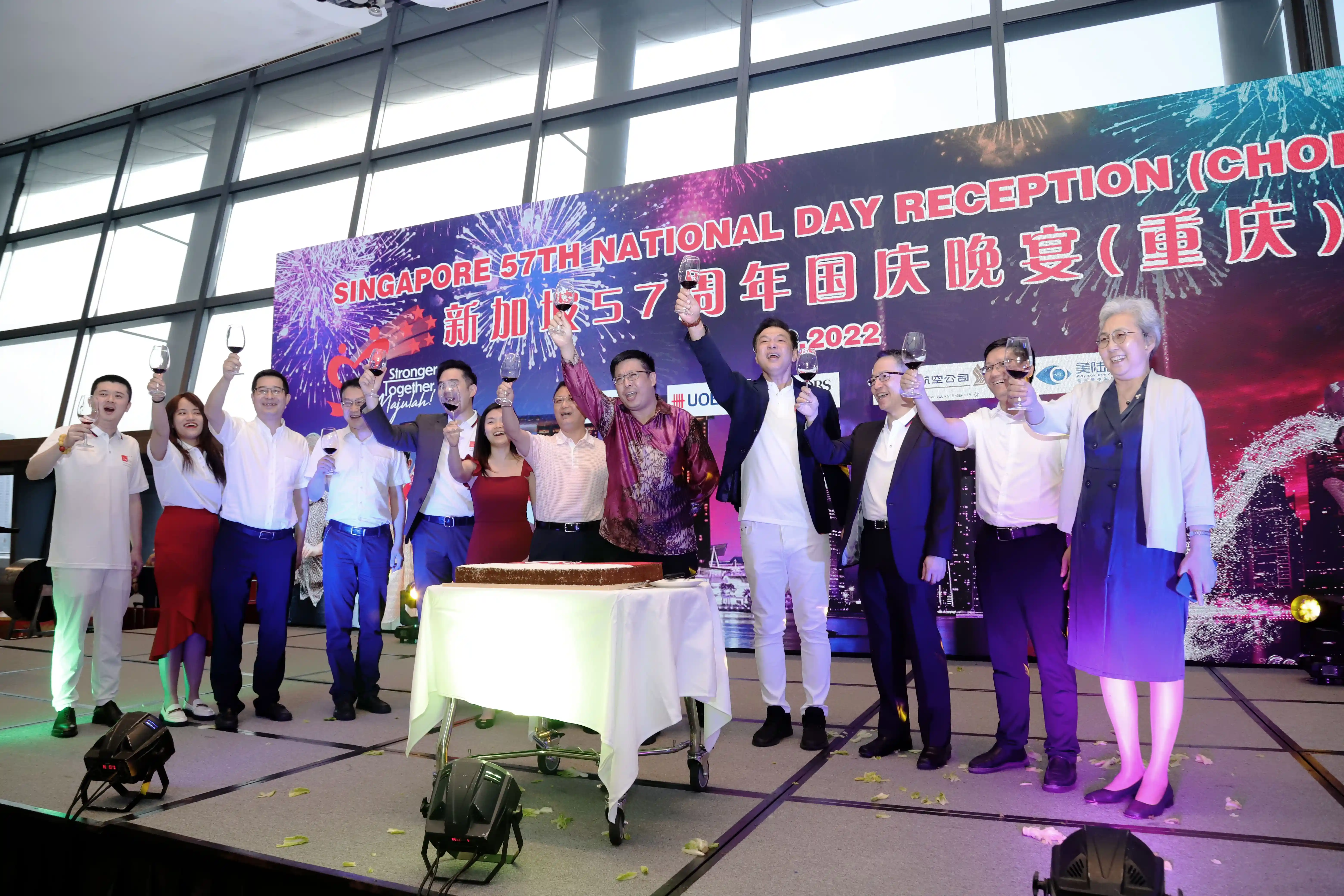 Group raises wine glasses on stage at Singapore's 57th National Day reception; cake on table, banner in background.