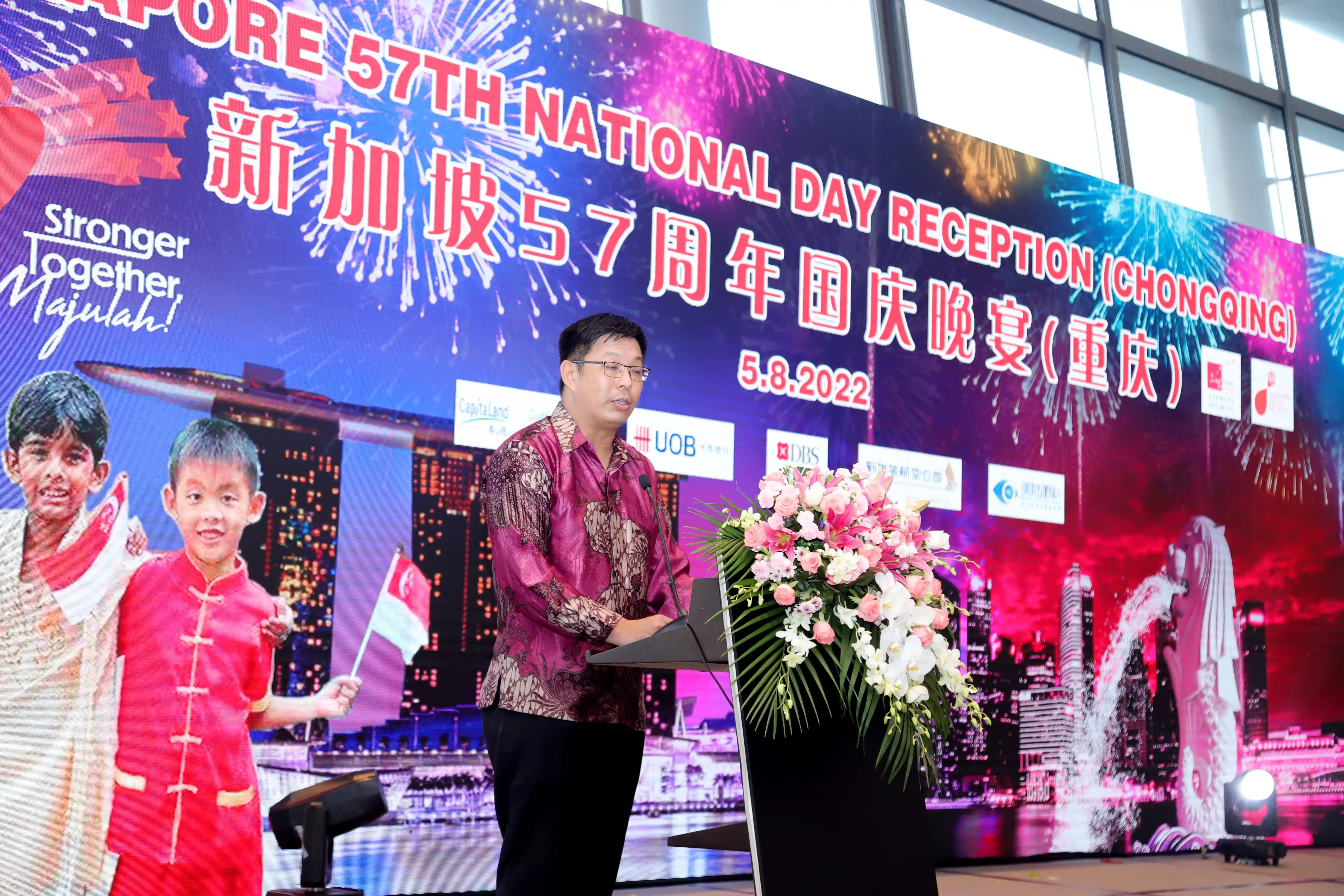 Person speaks at Singapore 57th National Day event, with floral arrangement and branded backdrop.