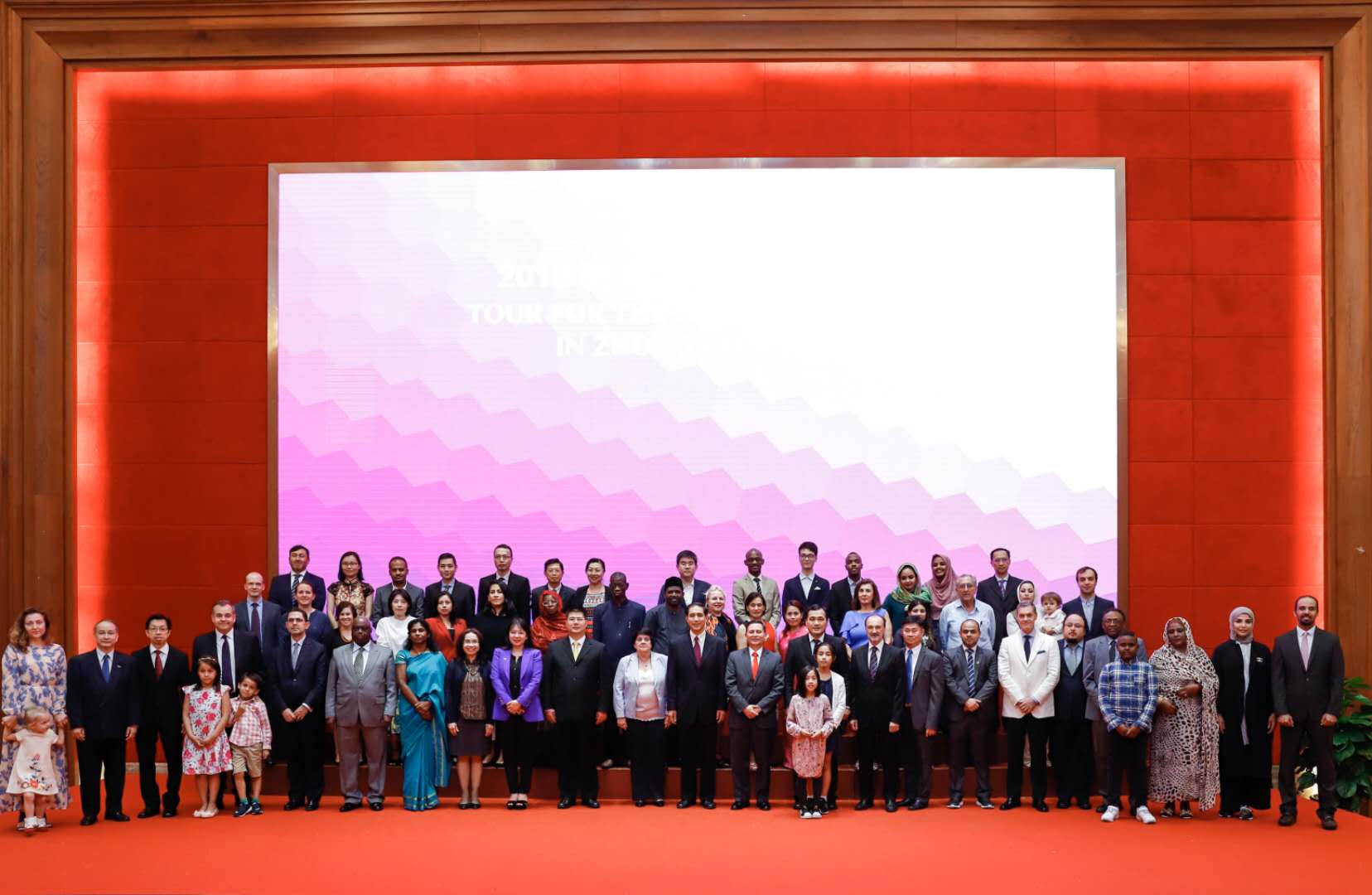 Group of people posing in front of large screen on red carpet, wood paneled wall.