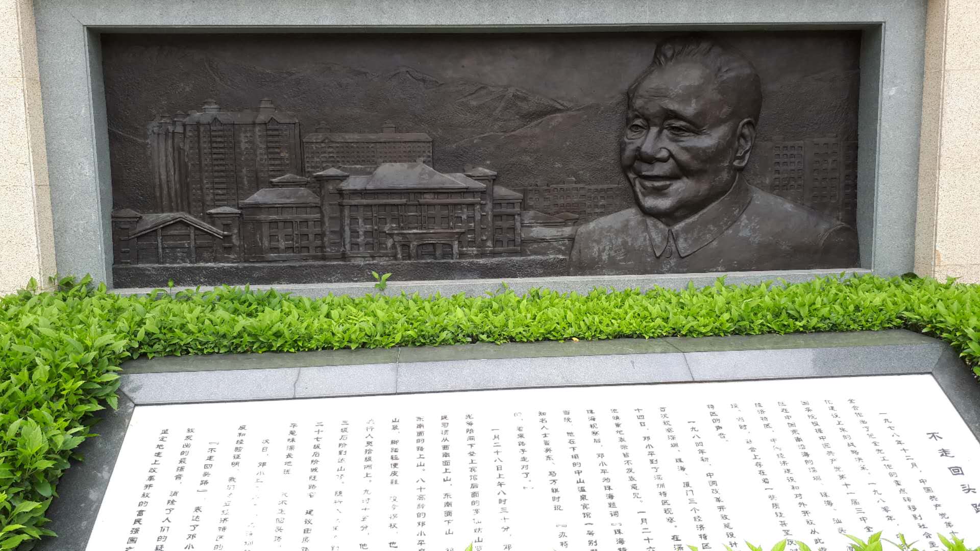 Bronze relief of Deng Xiaoping next to buildings, with Chinese text on stone.