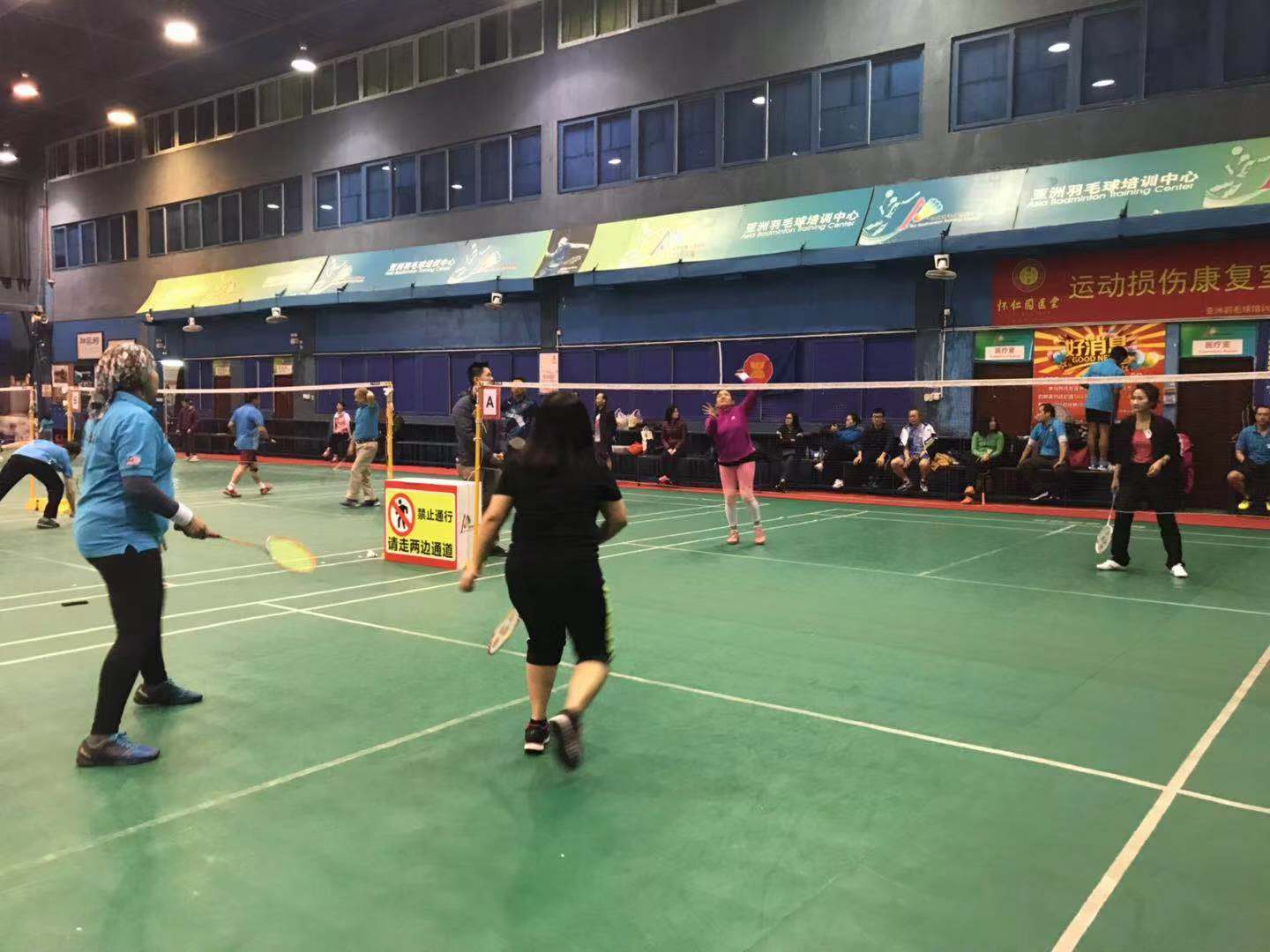 People playing badminton on a green court in a large indoor gymnasium.