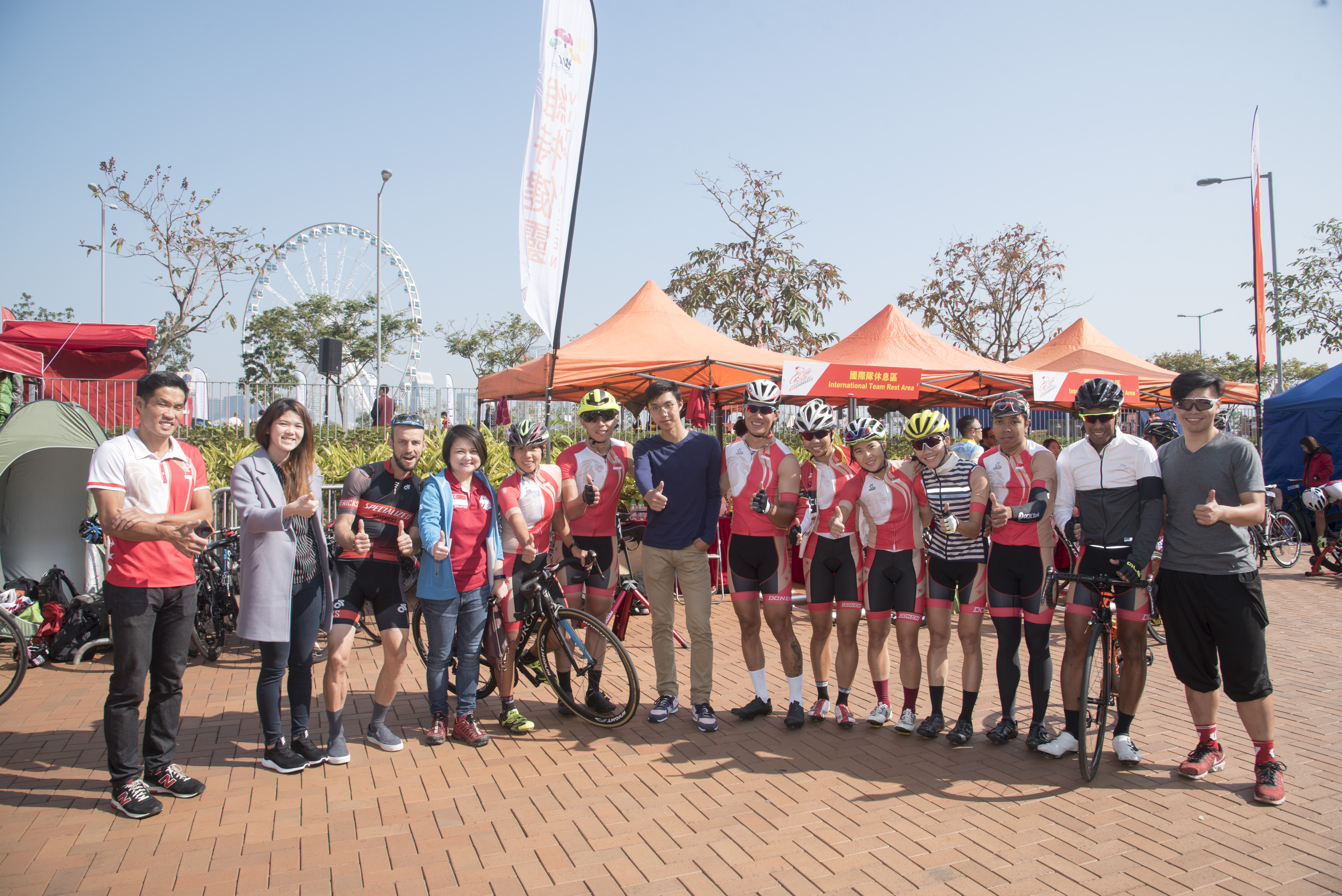 Group of cyclists in red and white gear giving thumbs up, with bikes and tents in the background.