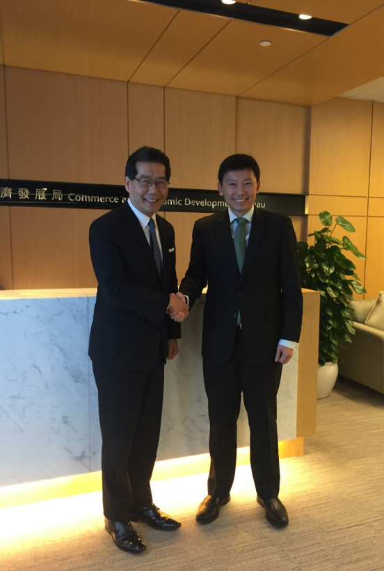Two men in suits shake hands in front of a "Commerce and Economic Development" sign.