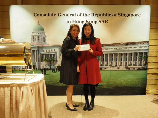 Two women hold a check in front of a "Consulate-General of Singapore" backdrop.
