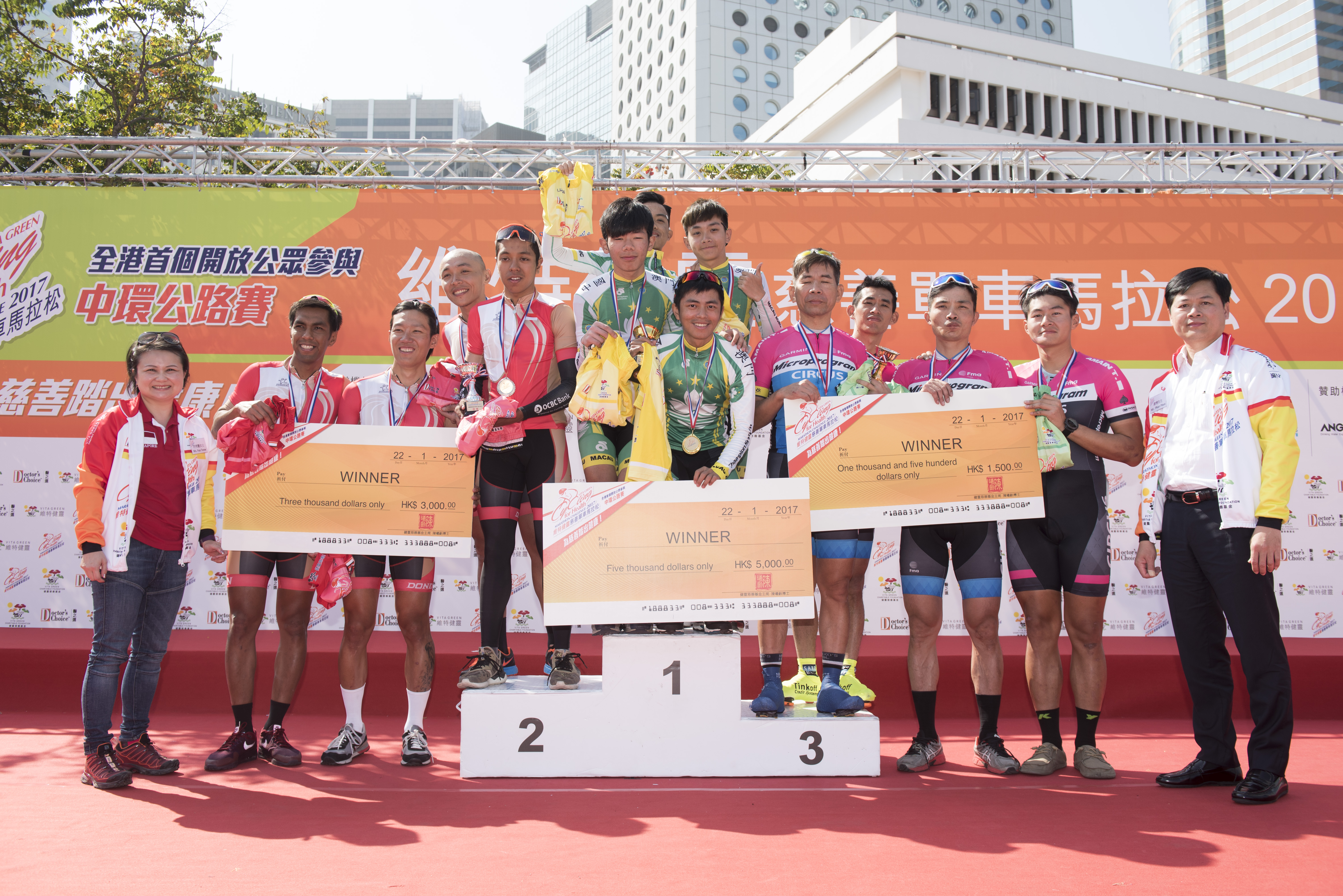Cyclists on a podium holding checks after a race, with a backdrop banner.