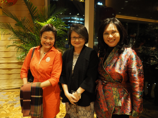 Three Asian women pose, indoors, wearing different colorful outfits in front of a green plant.
