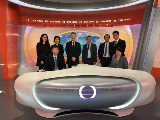 Eight people in business attire in a TVB News studio with a curved white desk.