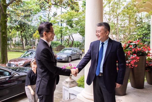 Two men in suits exchanging a gift bag in front of a mountain painting.
