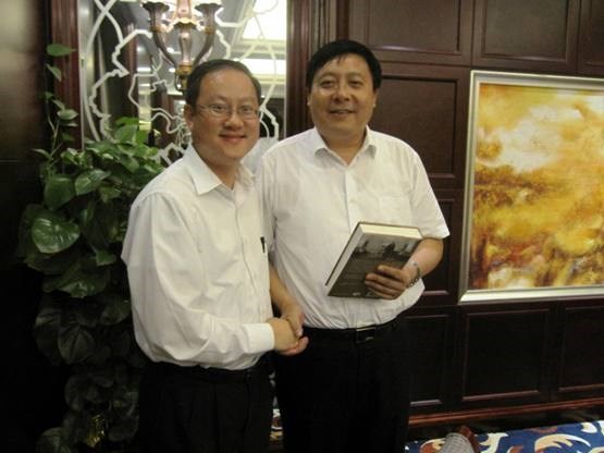 Two men in white shirts shake hands; one holds a book. Abstract art behind them.