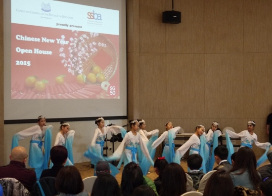 Young dancers in white and blue costumes perform before an audience. A projector showing a slide.