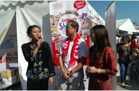 Three people stand before a "Singapore Fiesta" banner; one speaks into a microphone.