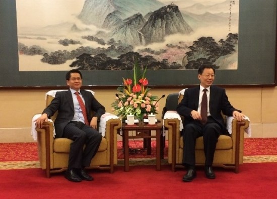 Two men in suits sit in armchairs with a table between them. A landscape painting hangs behind them.