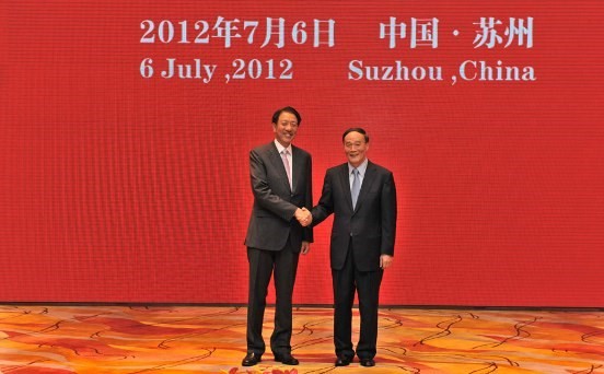 Two men in suits shaking hands in front of a red backdrop with the text "2012年7月6日 中国·苏州 6 July ,2012 Suzhou, China"