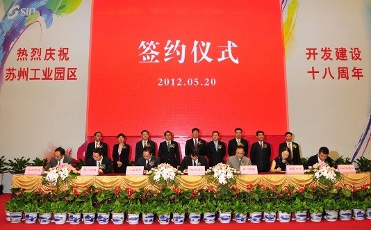 People in suits at a signing ceremony with banners and floral arrangements.