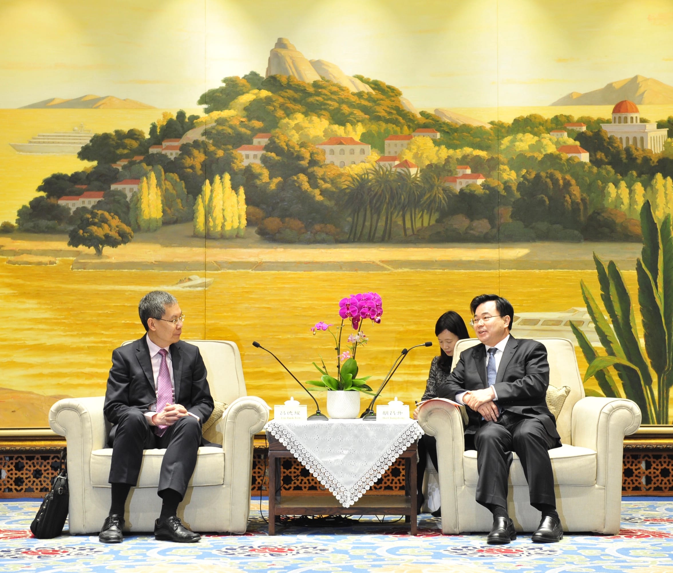 Two men in suits seated, microphones on table with orchids, mural behind.