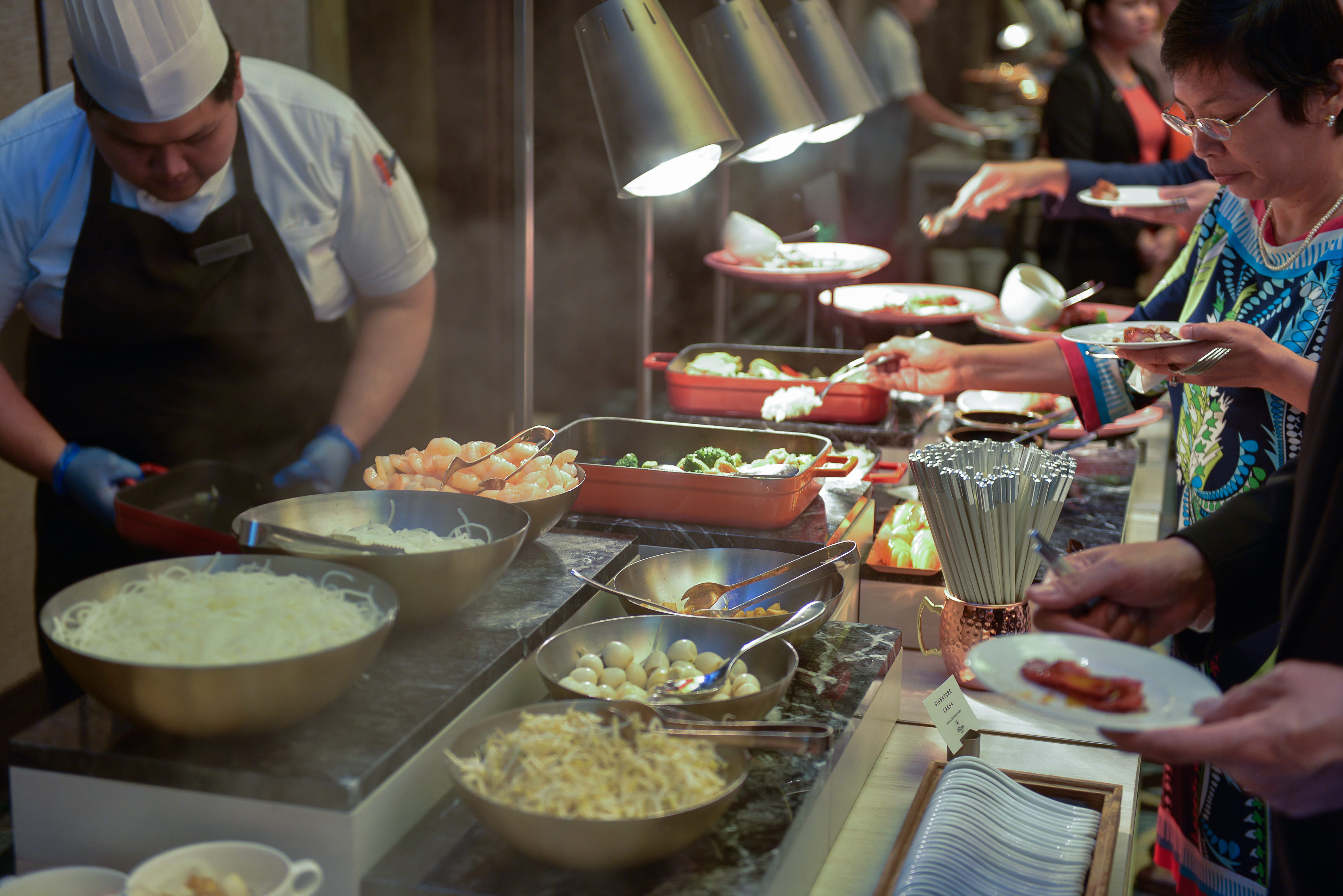 Seven women stand in a group, wearing casual and traditional clothing in front of floral decor.