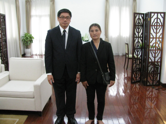 Two people stand indoors on a wooden floor. A man in a suit and a woman in a blazer with bare feet.