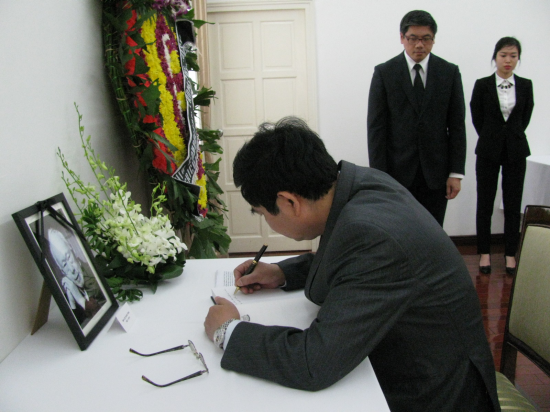 Man signs guest book near portrait, flowers, wreath. Two people stand in dark suits.