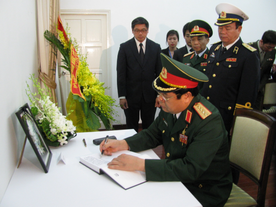 Military personnel in uniform sign a book near flowers and a framed photo on a white table.