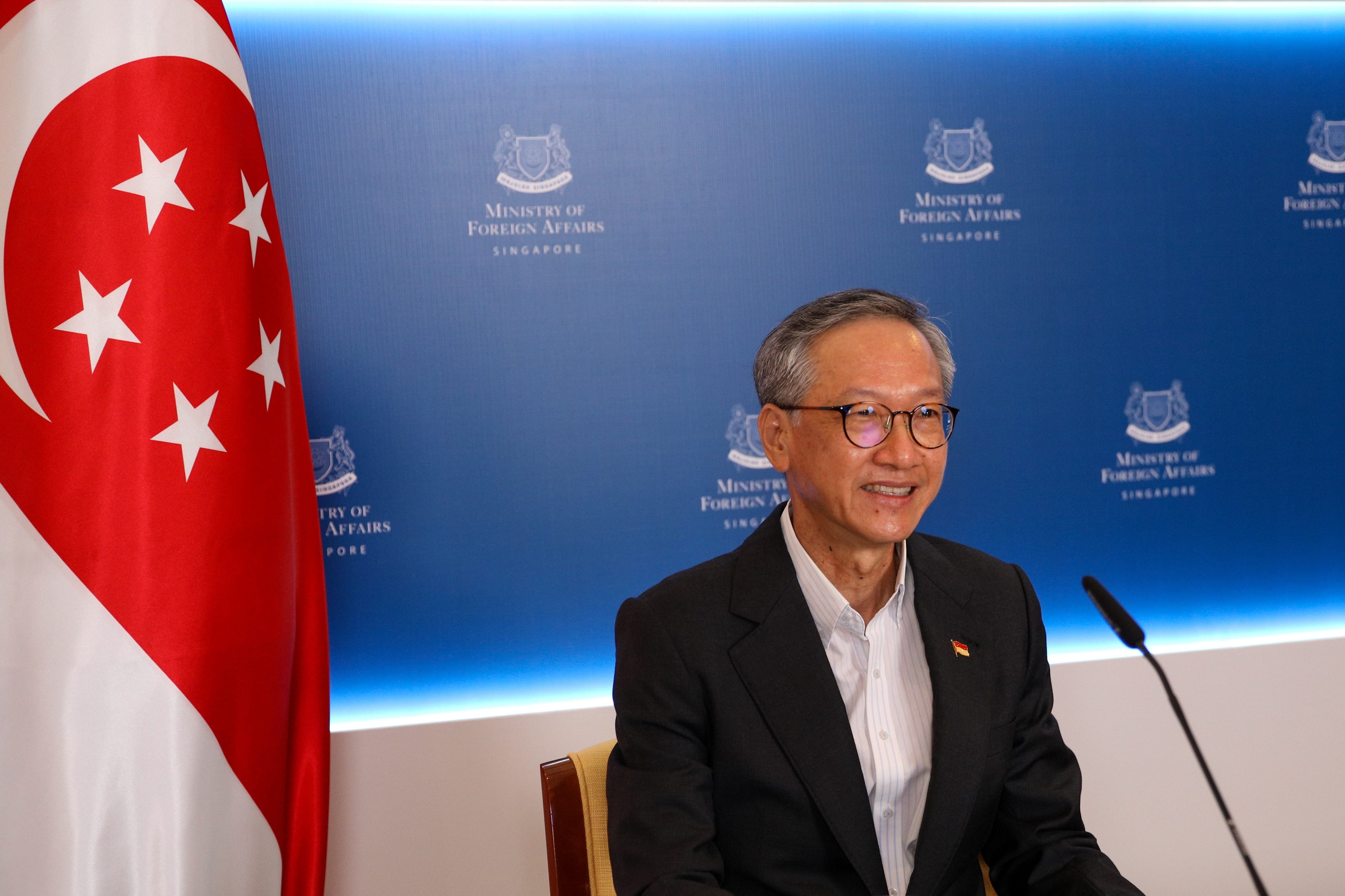 Man in suit with Singapore flag, speaking in front of Ministry of Foreign Affairs backdrop.