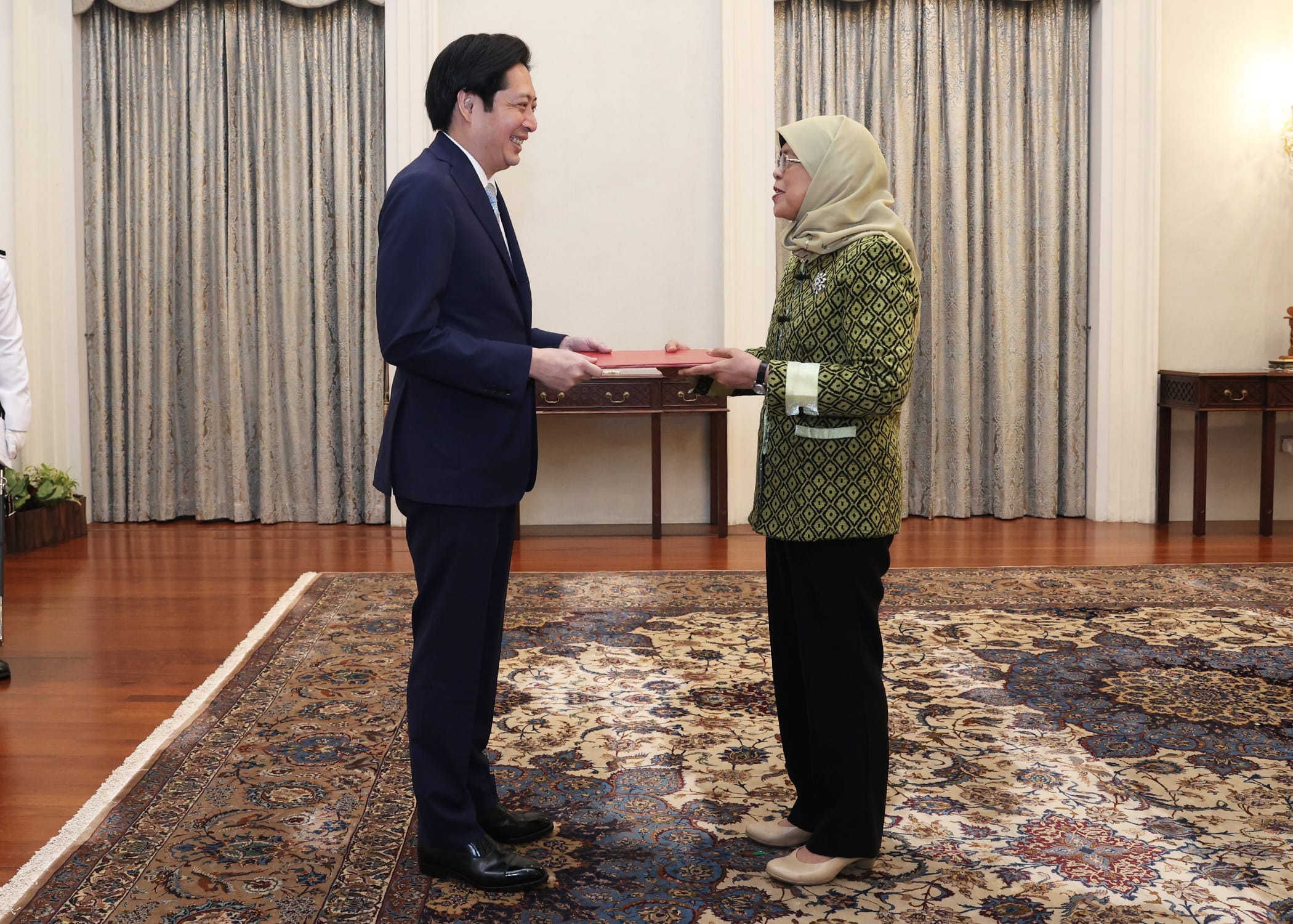 Man in suit hands red folder to woman in hijab, indoors on patterned carpet.