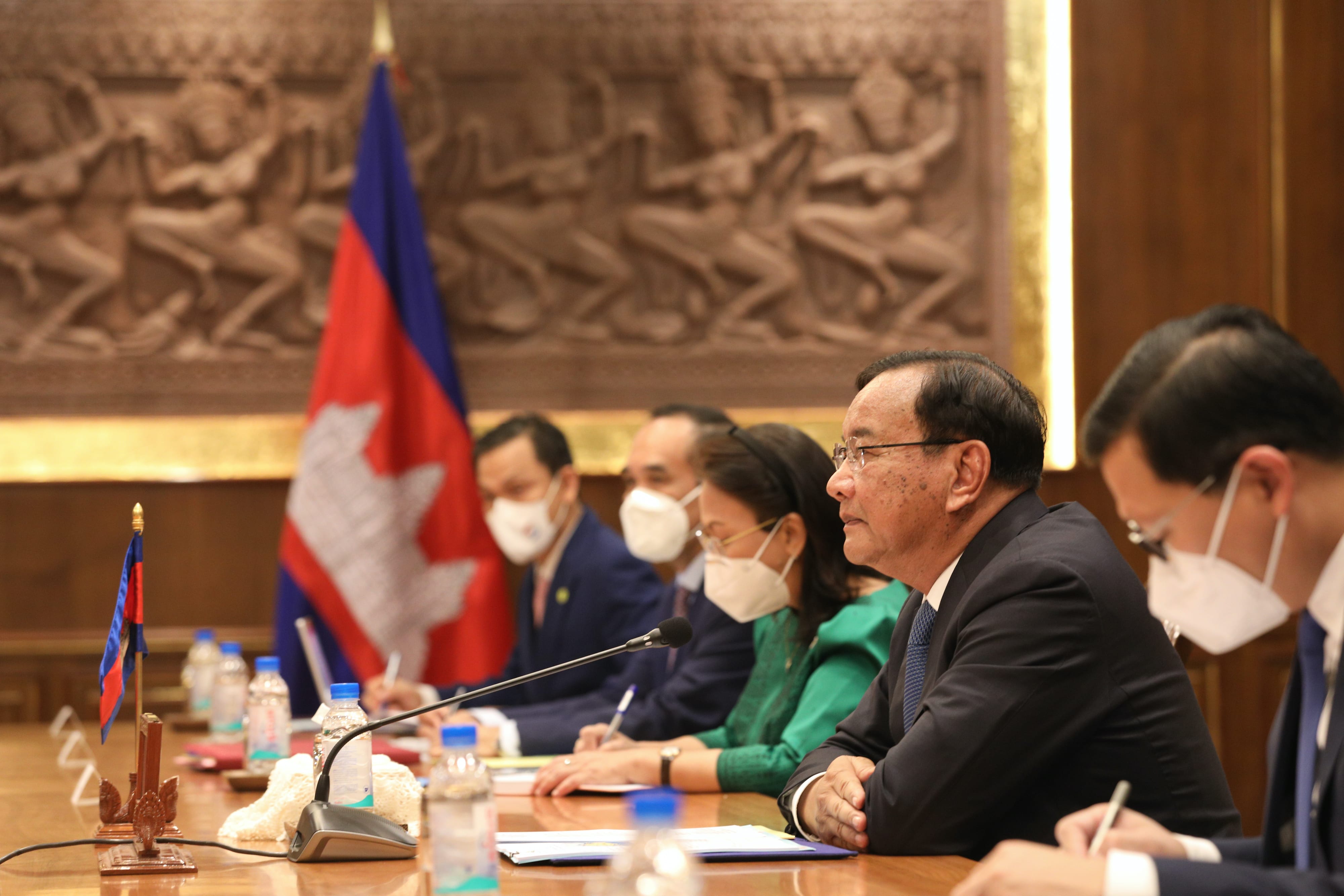 People at conference table with Cambodian flags, microphones, water bottles, and carved backdrop.