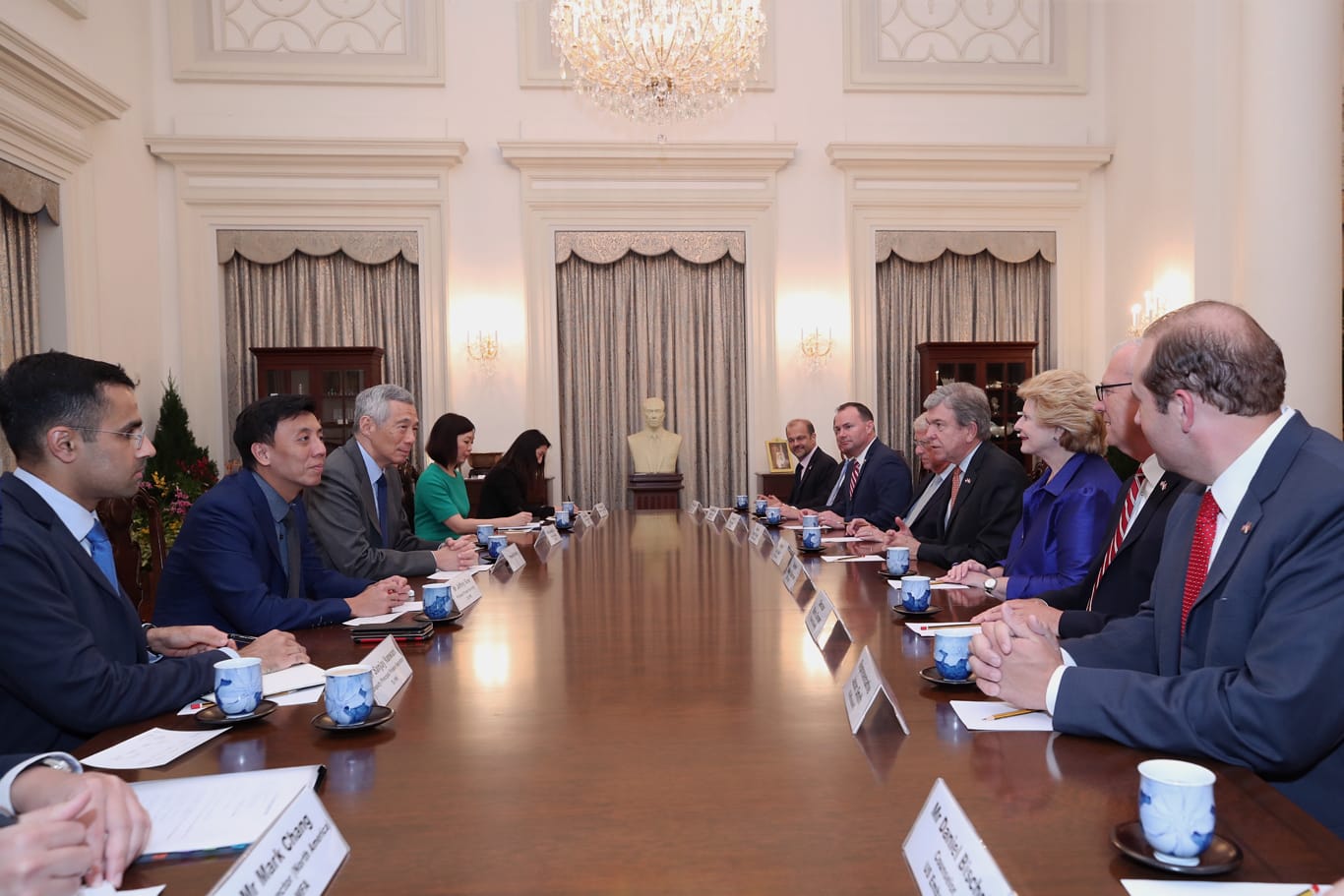 People at long conference table, with name cards and teacups, under a chandelier. Bust in background.
