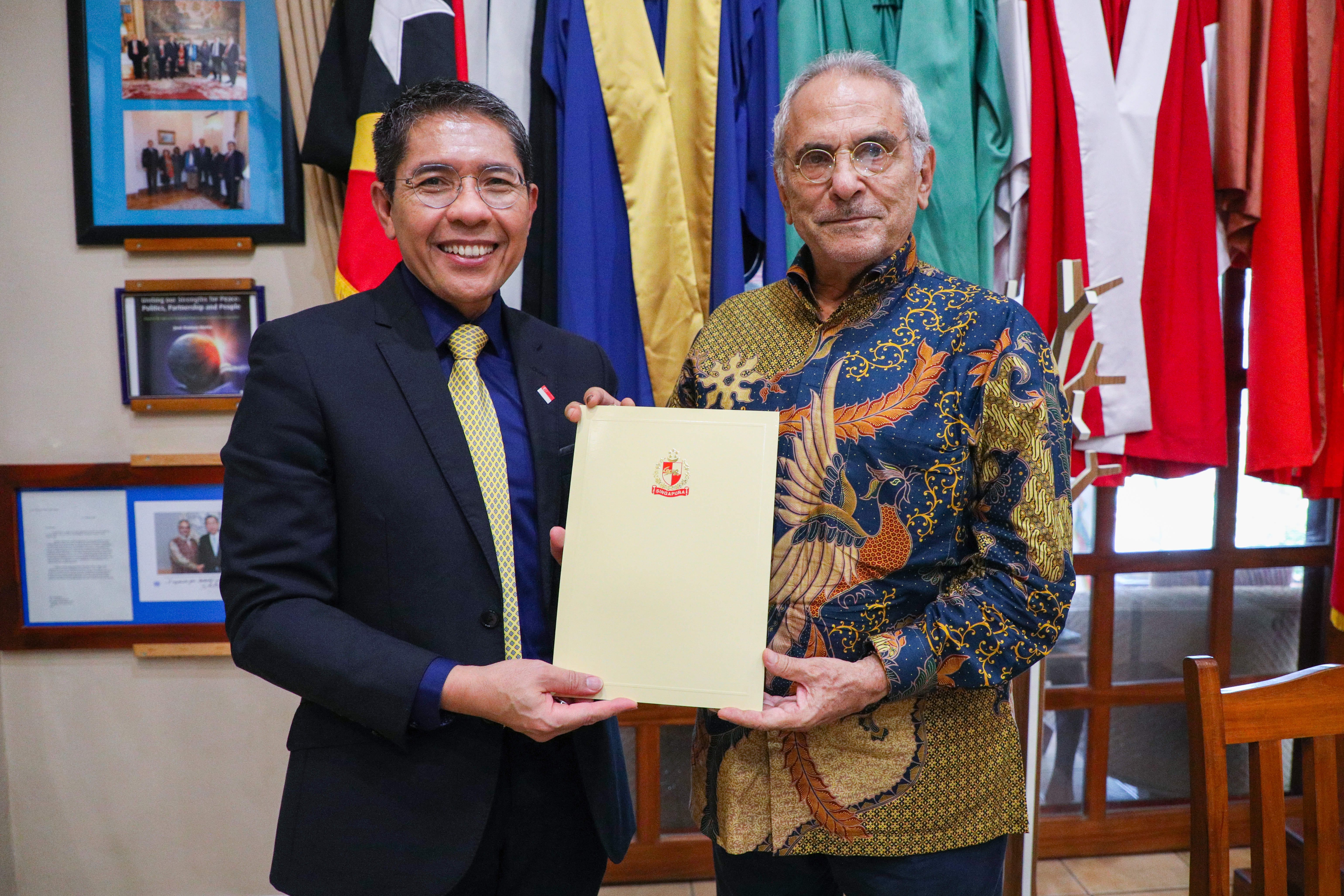 Two men hold a document with Singapore coat of arms, wearing a suit and patterned shirt.