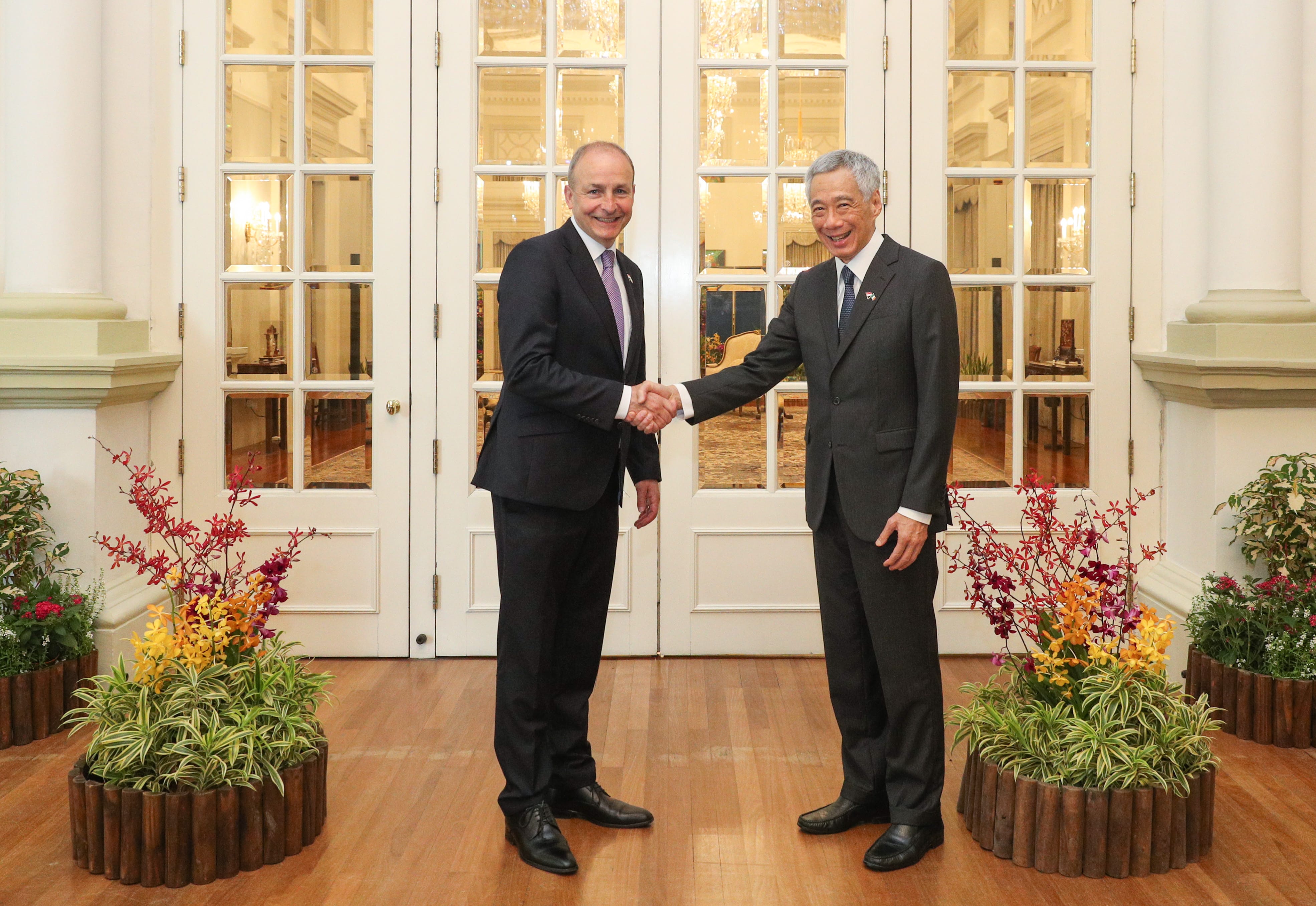 Two men in suits shaking hands in a room with wooden floors, a rug, table, and patterned curtains.