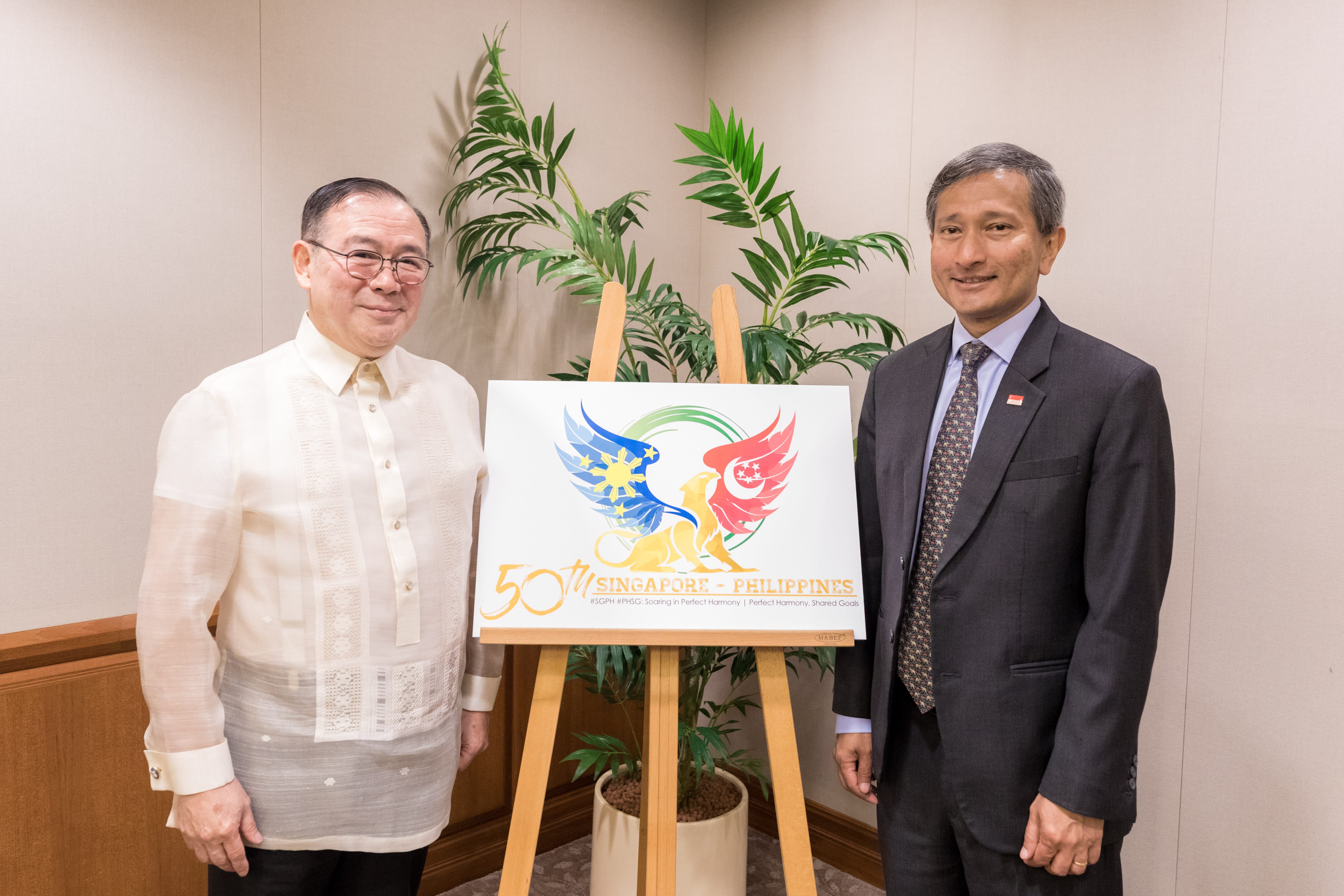 Two men flank an easel displaying "50th Singapore-Philippines" logo, one wears a suit, the other a traditional shirt.