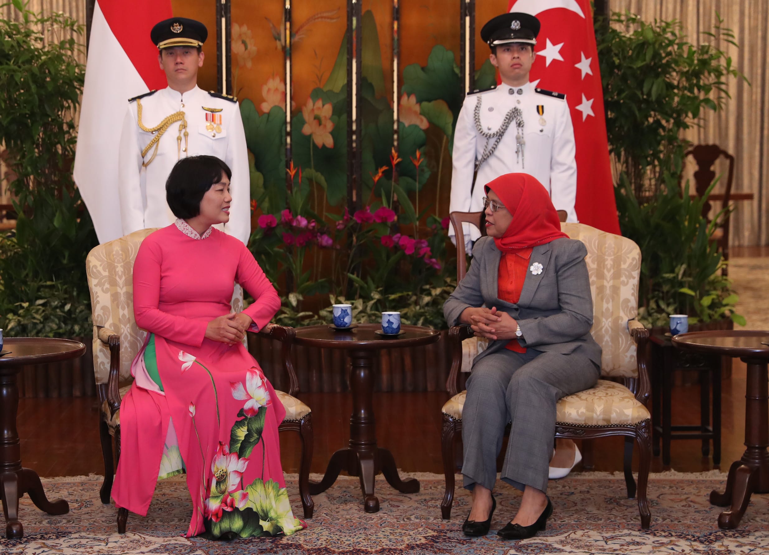 Halimah Yacob, wearing a red hijab and grey suit, sits across from a guest with guards behind them.