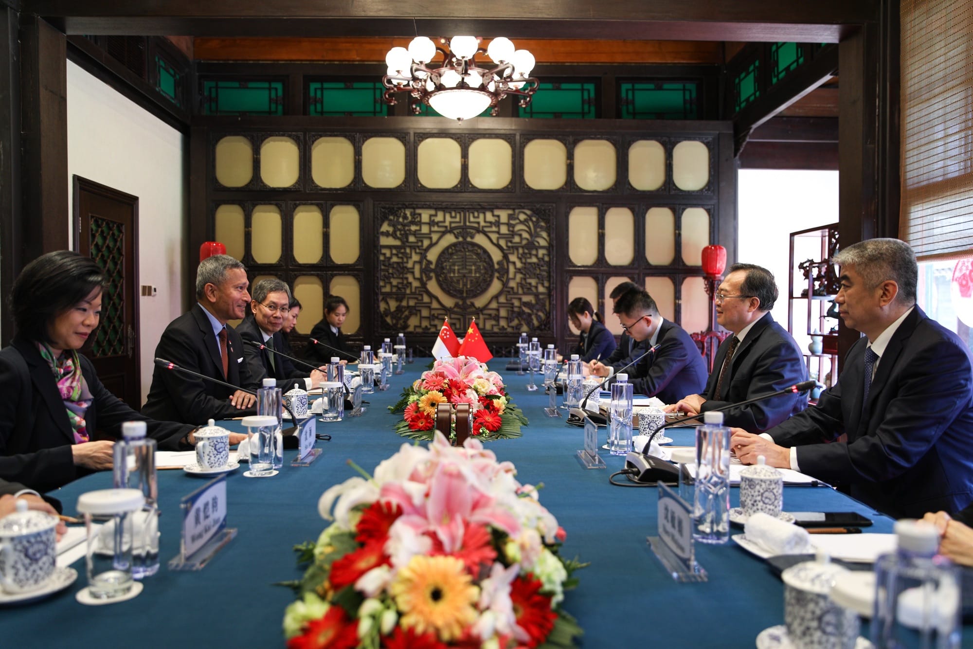 People seated at a long table with floral centerpiece, flags, water, and microphones.