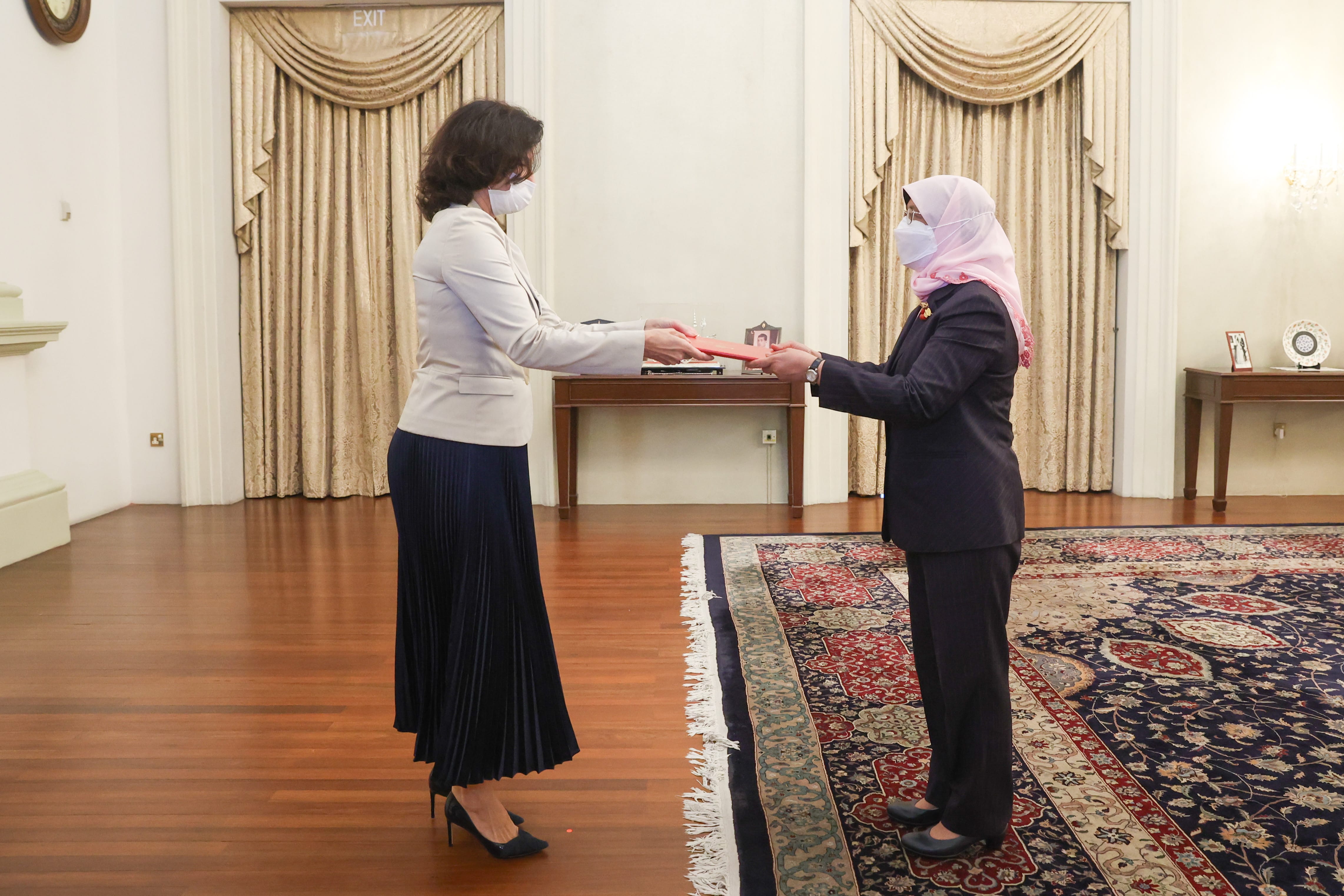 Two women in face masks exchange a red folder in an ornate room.
