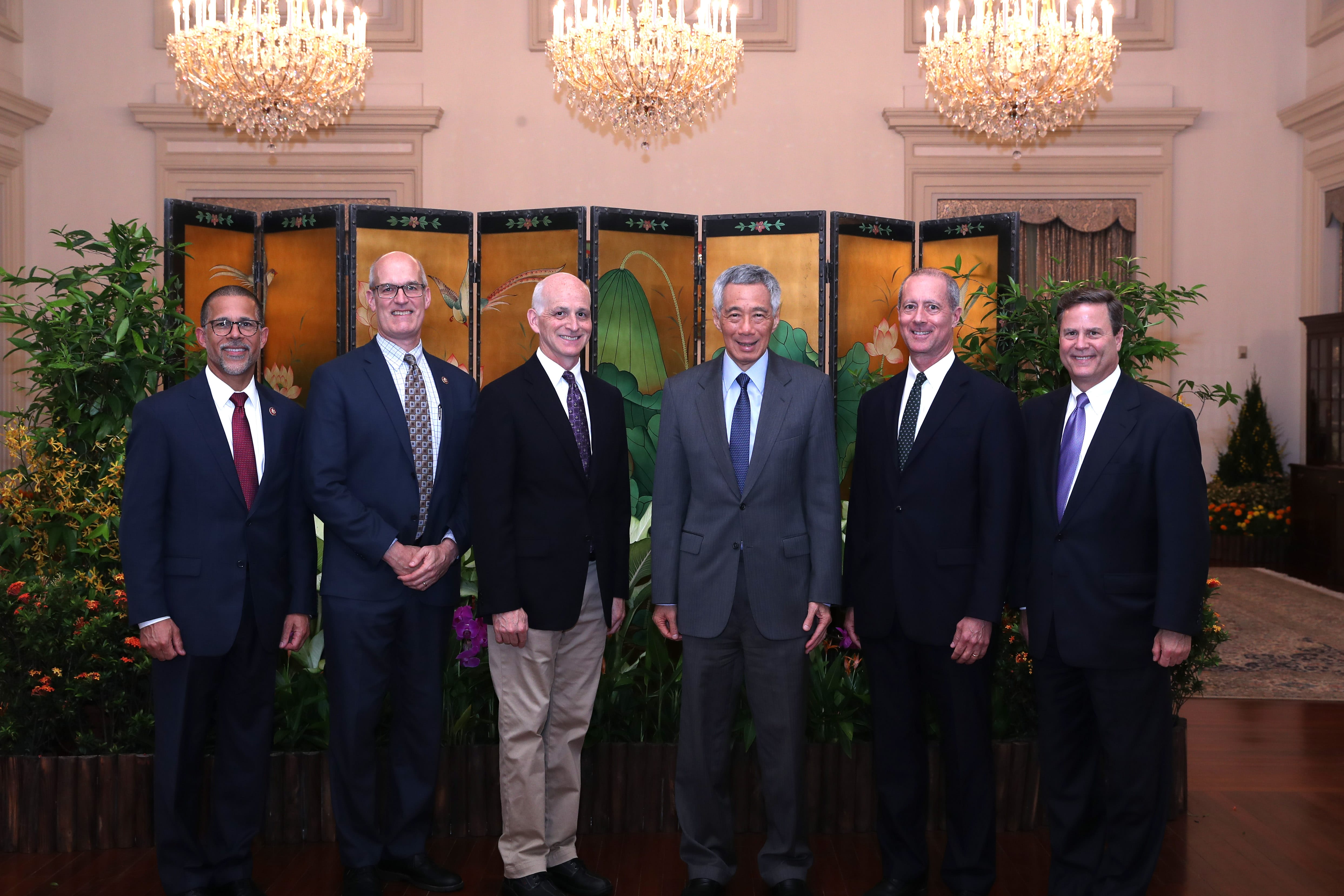 Six men in suits stand posed in front of an Asian-style screen and chandeliers.