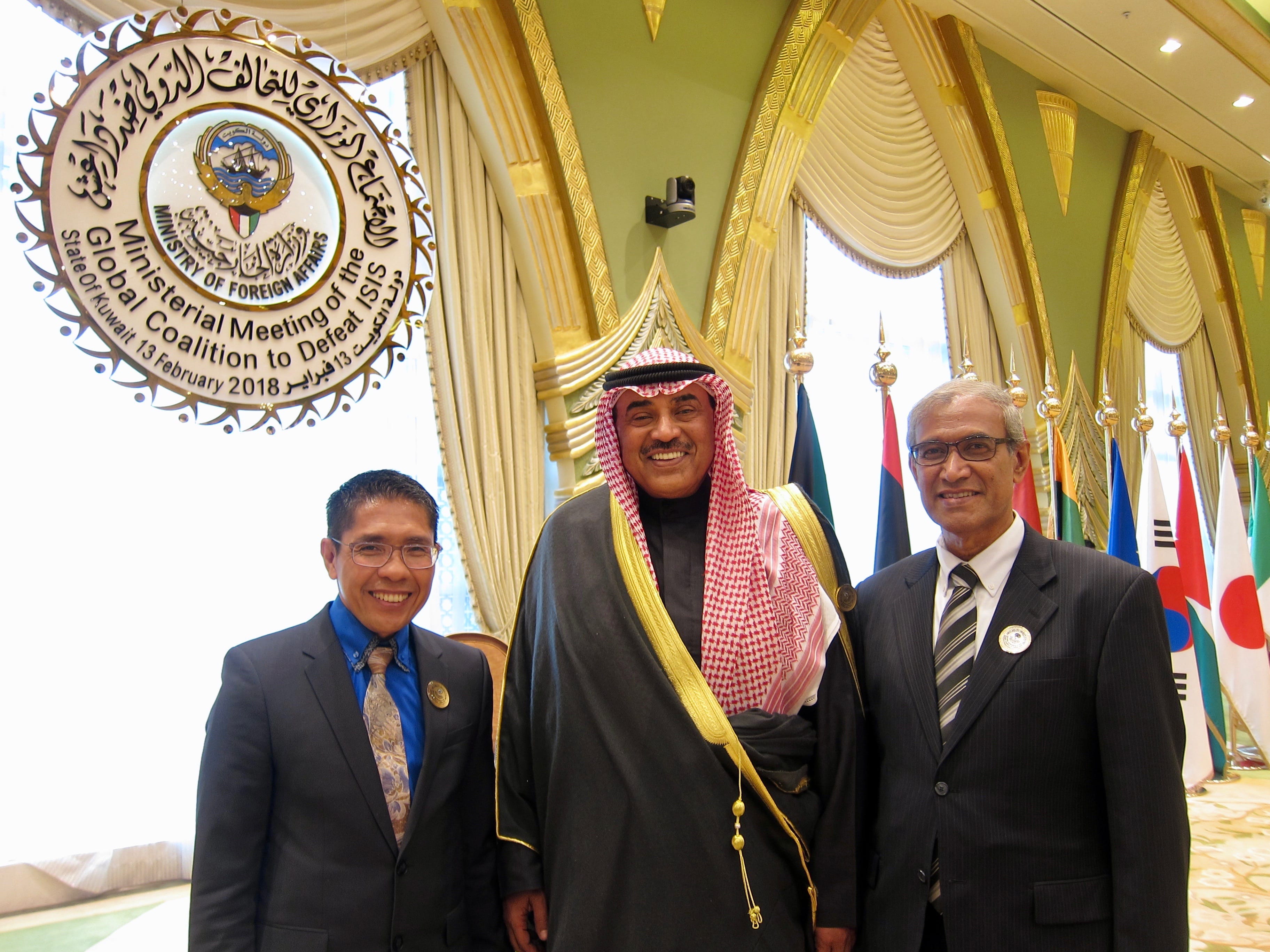 Three men in suits pose before flags and a "Global Coalition to Defeat ISIS" sign.