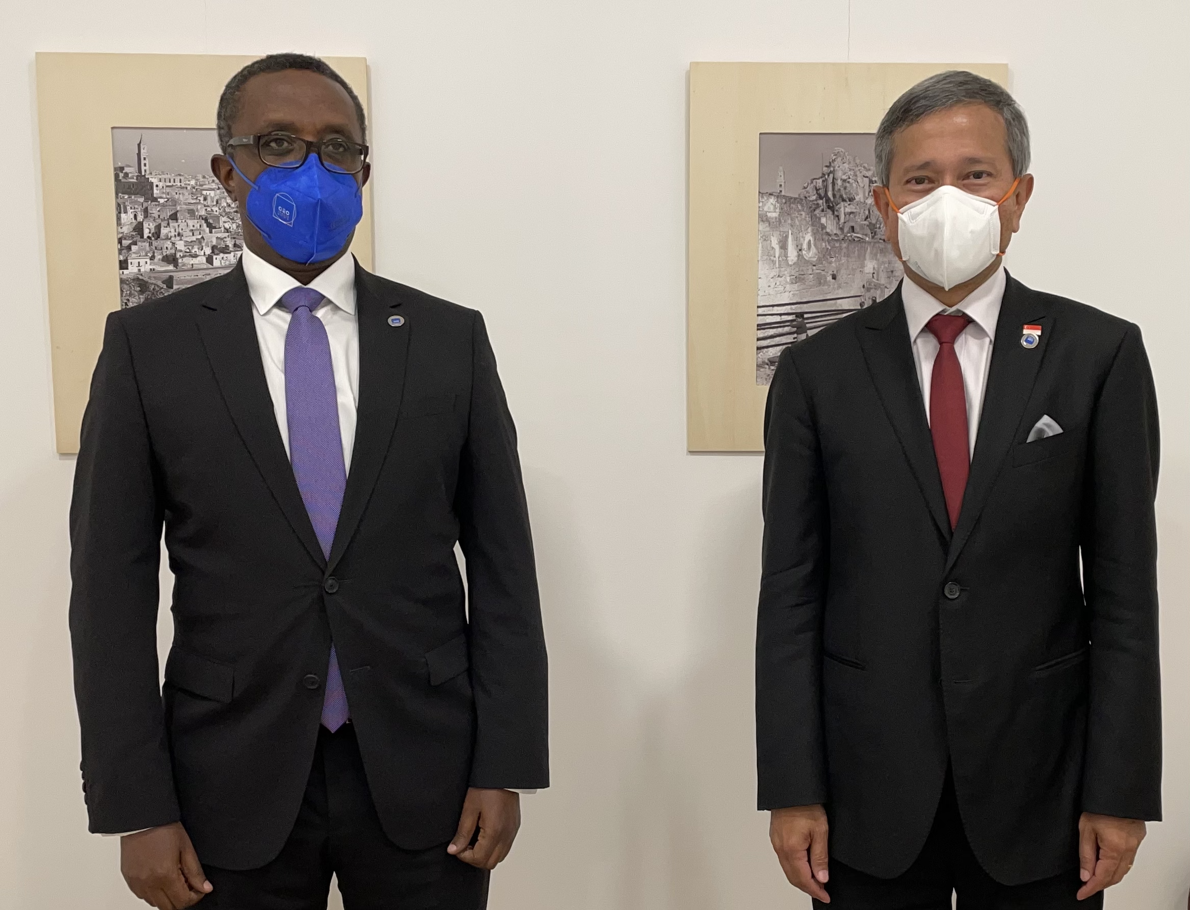 Two men in suits wear masks, standing in front of framed artwork on a white wall.