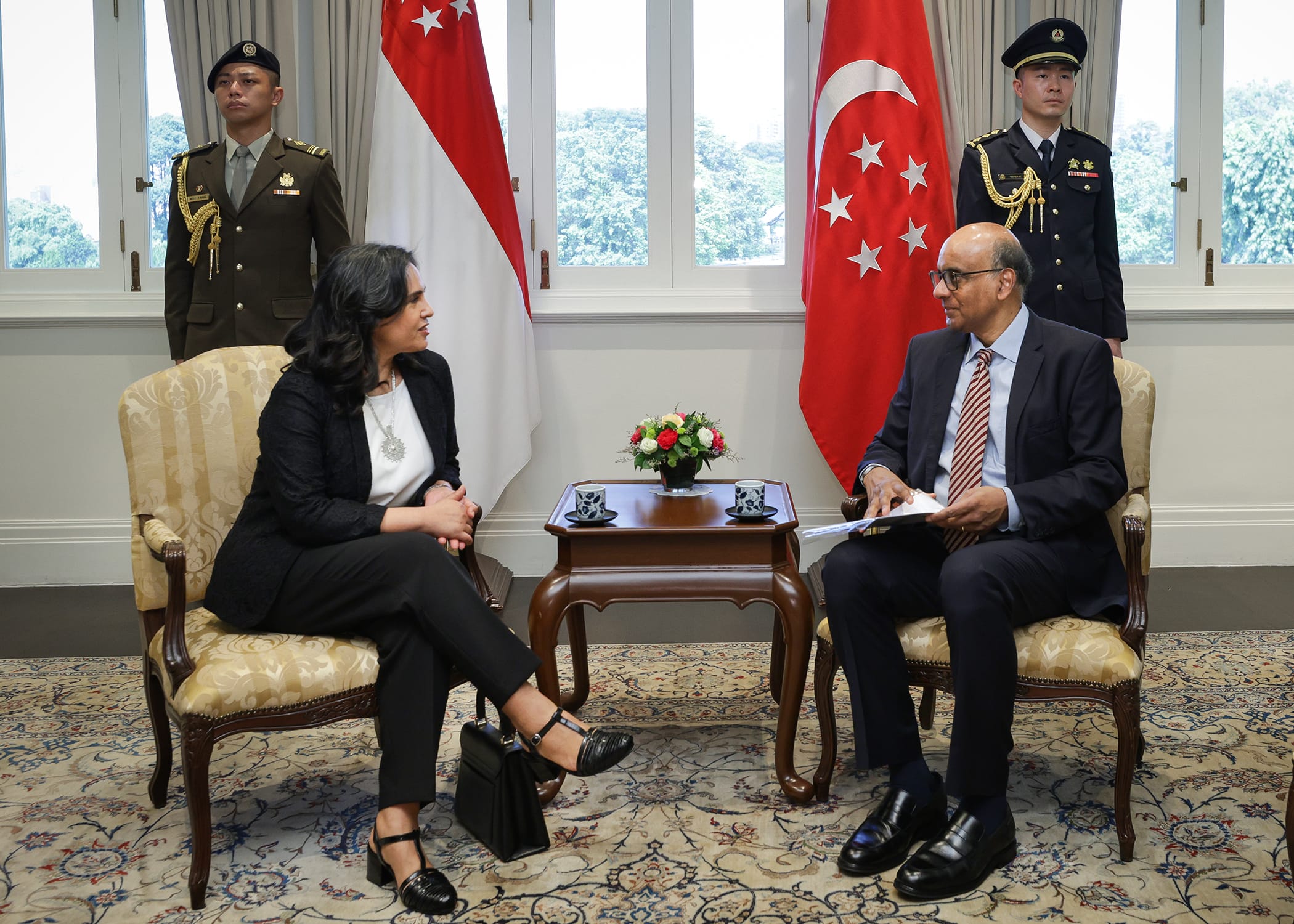 Man and woman seated in chairs, Singapore flags and guards behind them.