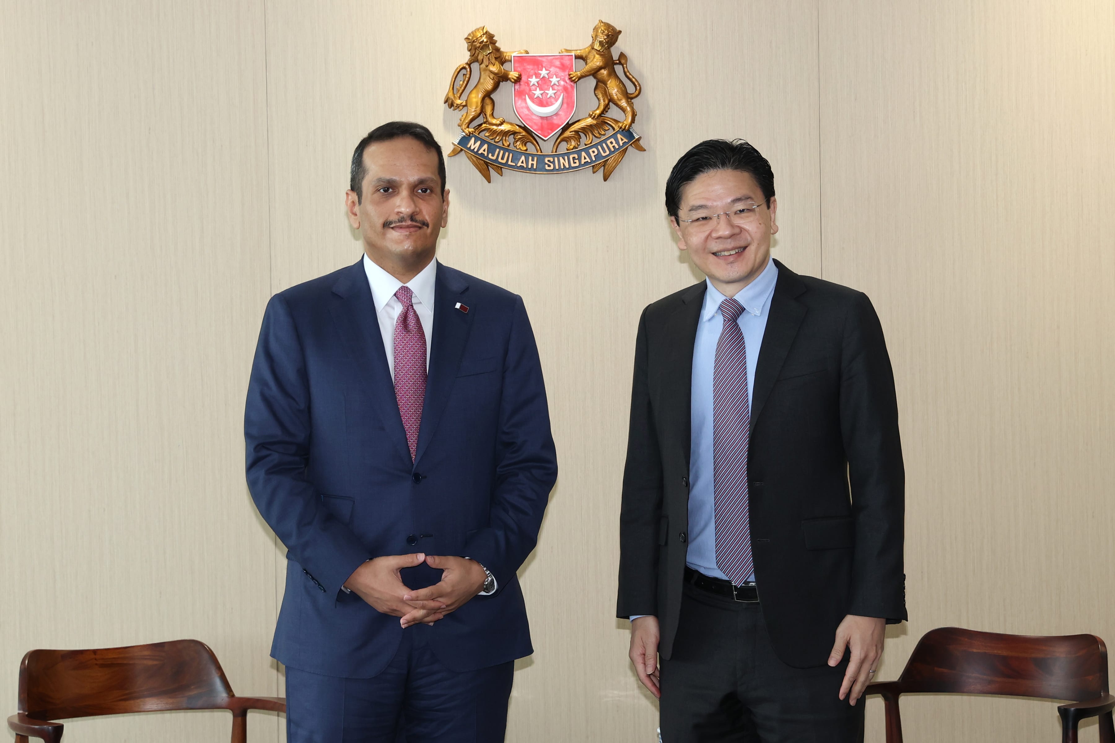 Two men in suits stand before the Singapore coat of arms on a light wall.