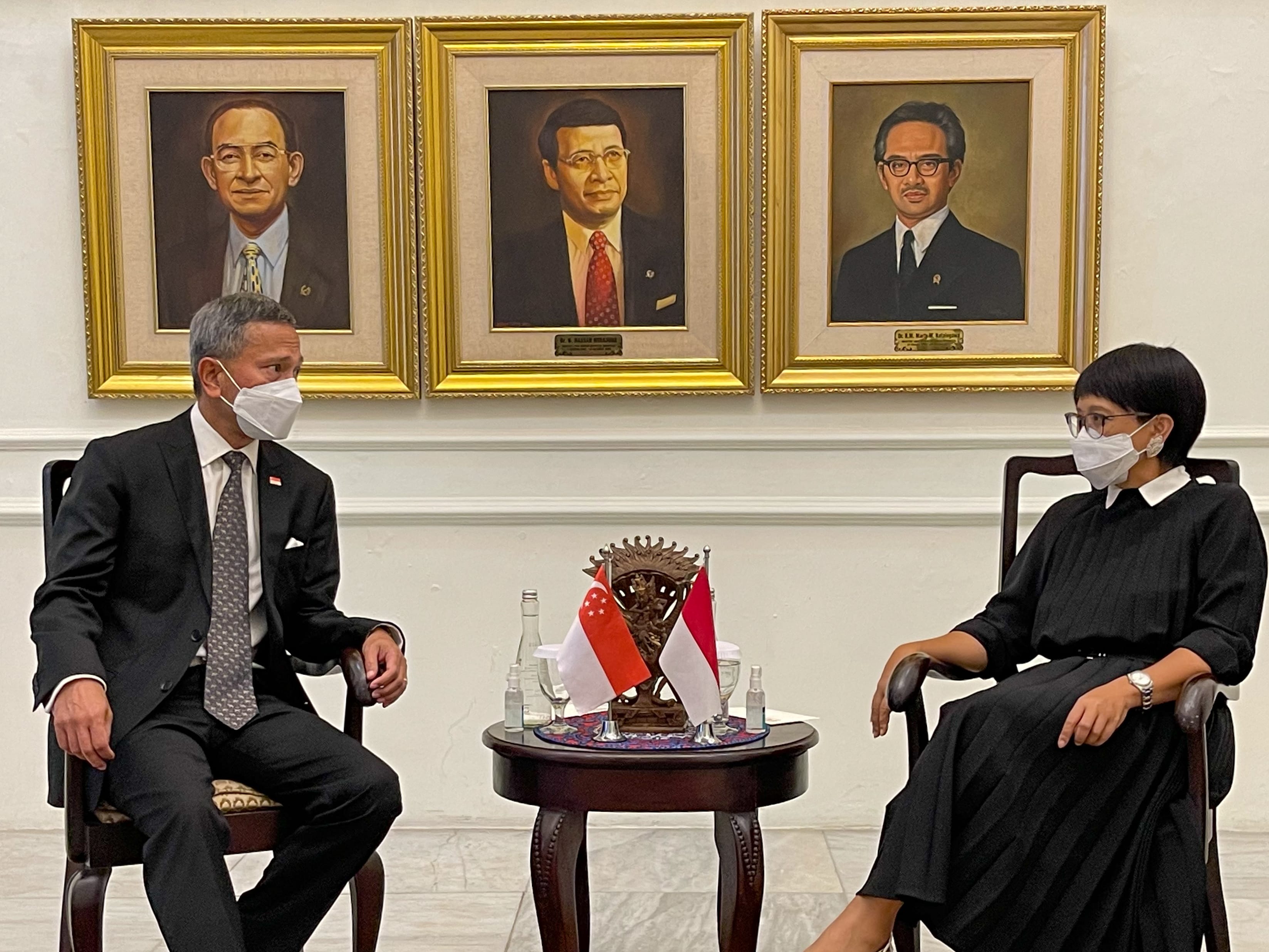 Two masked people in formal wear seated near table with flags of Singapore & Indonesia. Framed portraits on wall.
