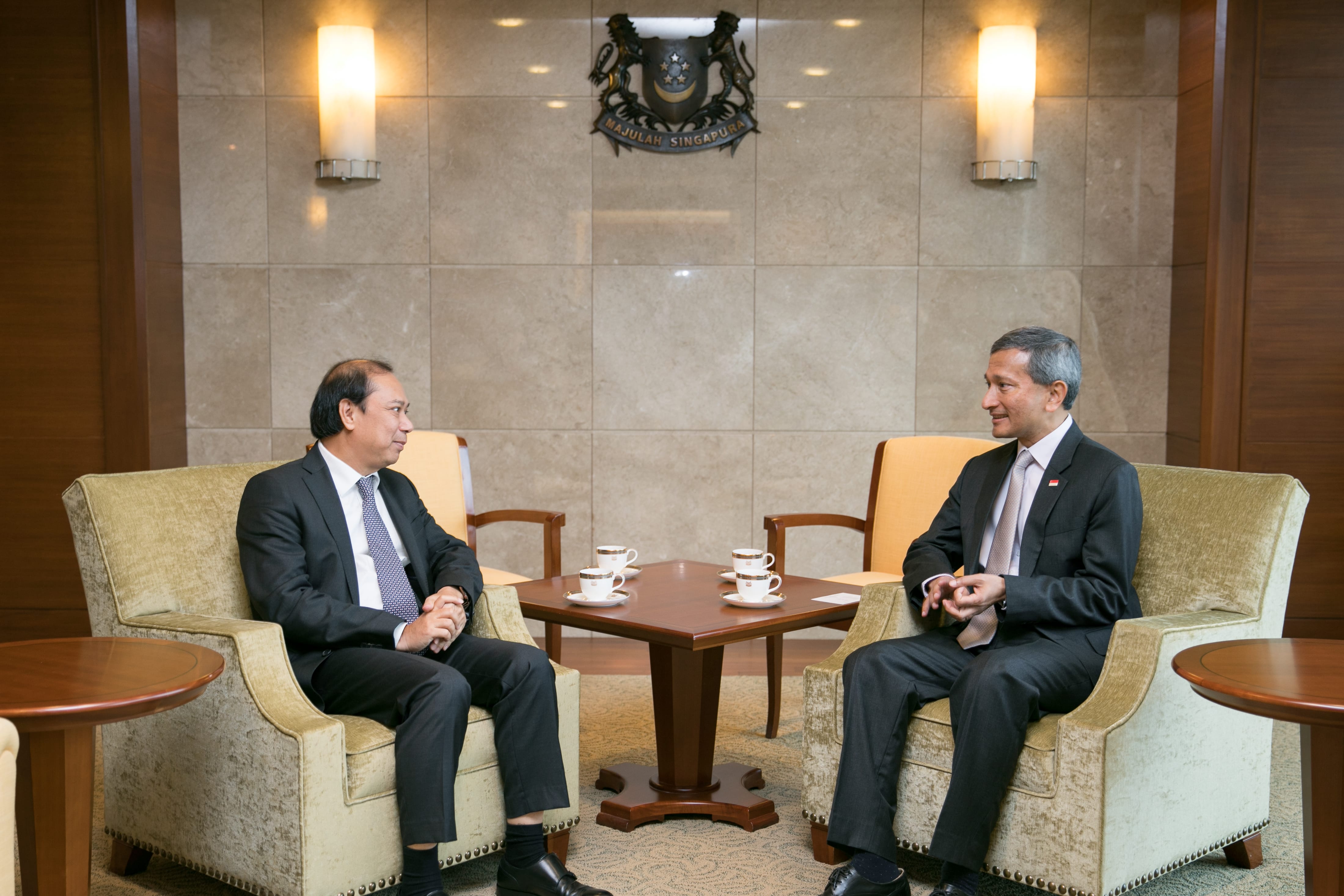 Two men in suits seated, facing each other with stacked teacups on table, Singapore coat of arms in background.