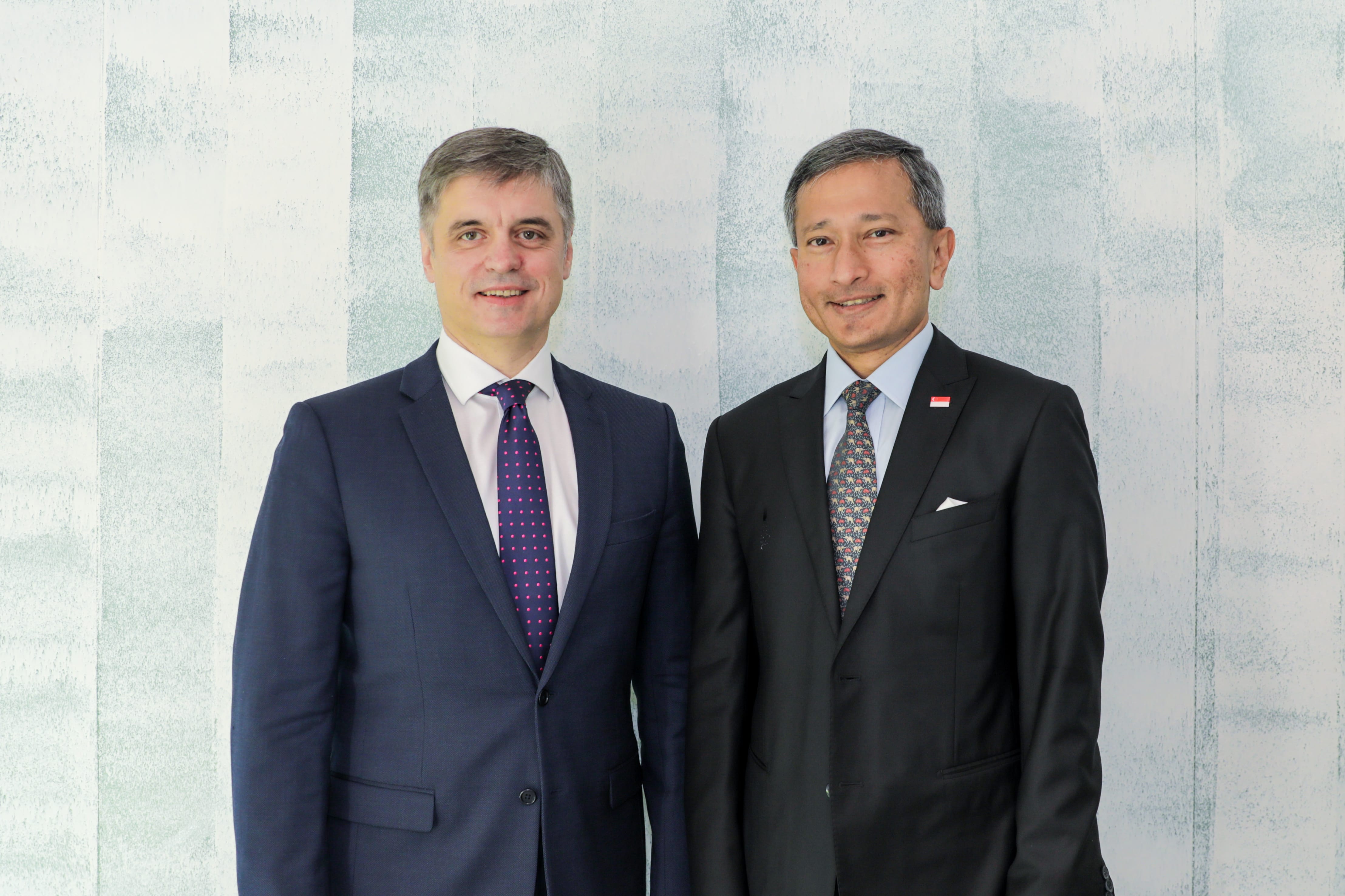 Two men in suits standing side-by-side against a textured white wall.
