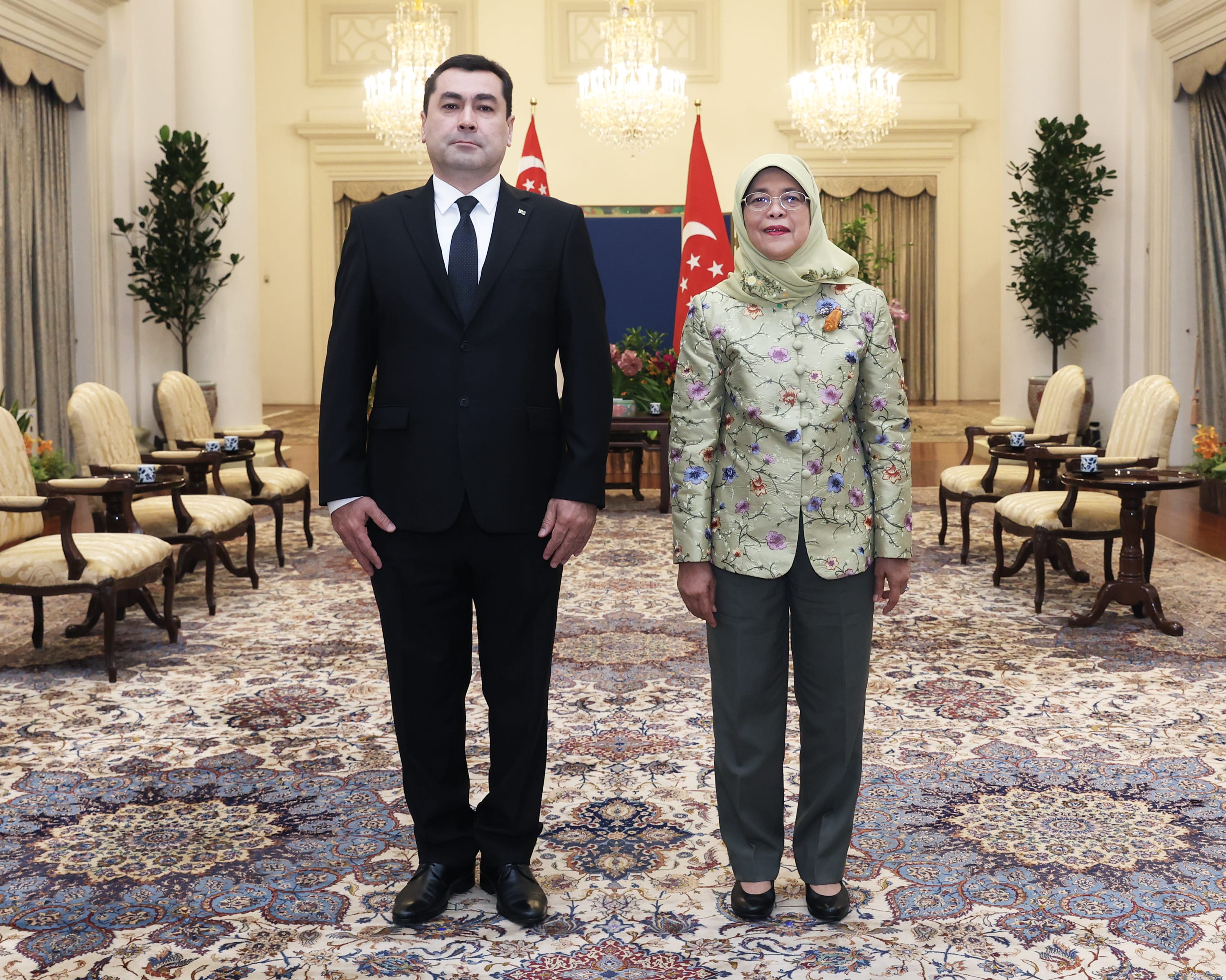 Two people stand in a room with Singapore flags, chandeliers, ornate carpet, and furniture.