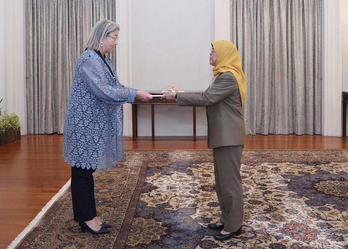 Two women exchange documents in a formal room with an ornate rug.