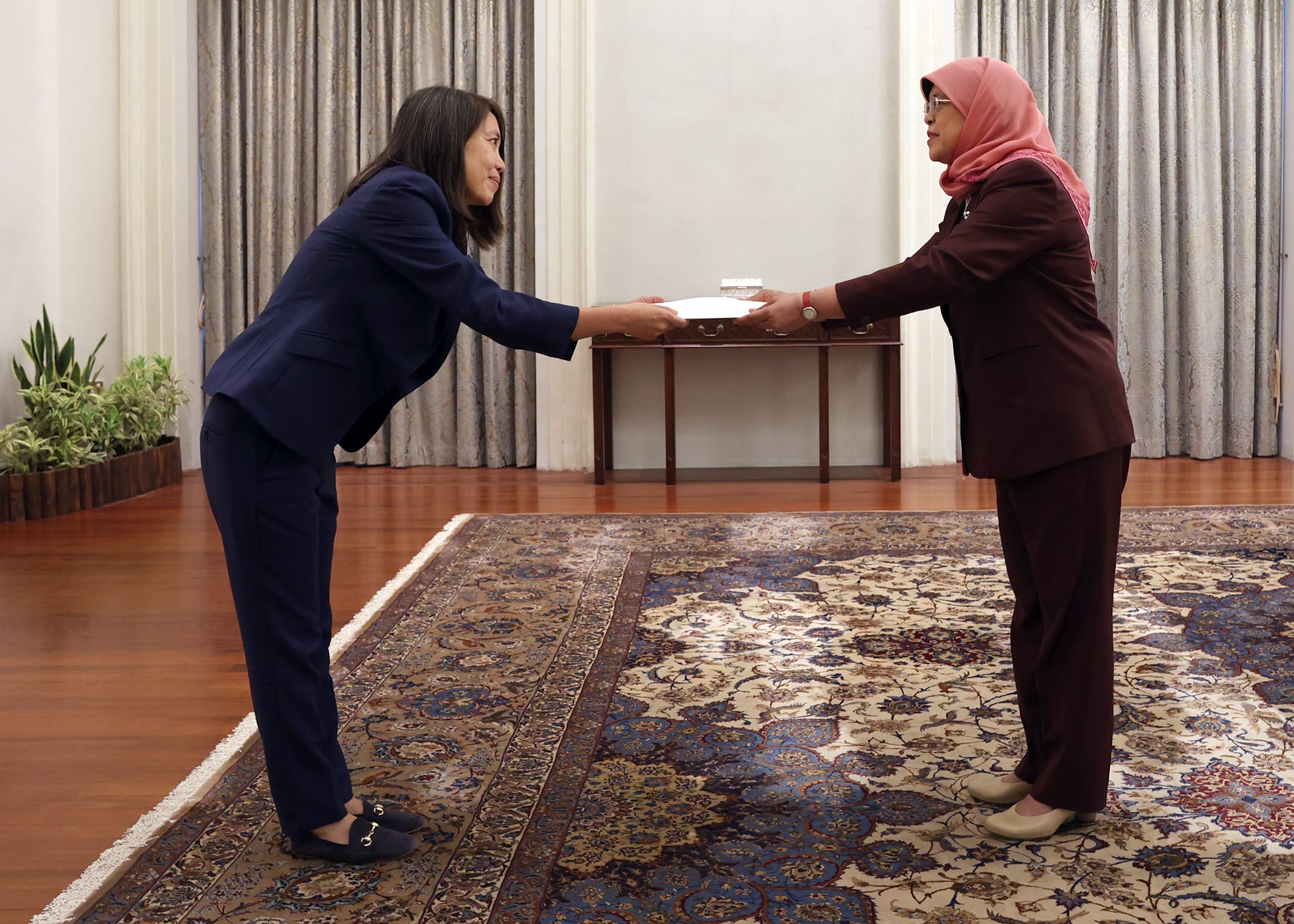 Two women in suits exchanging documents over a table on an ornate rug. One wears a pink hijab.