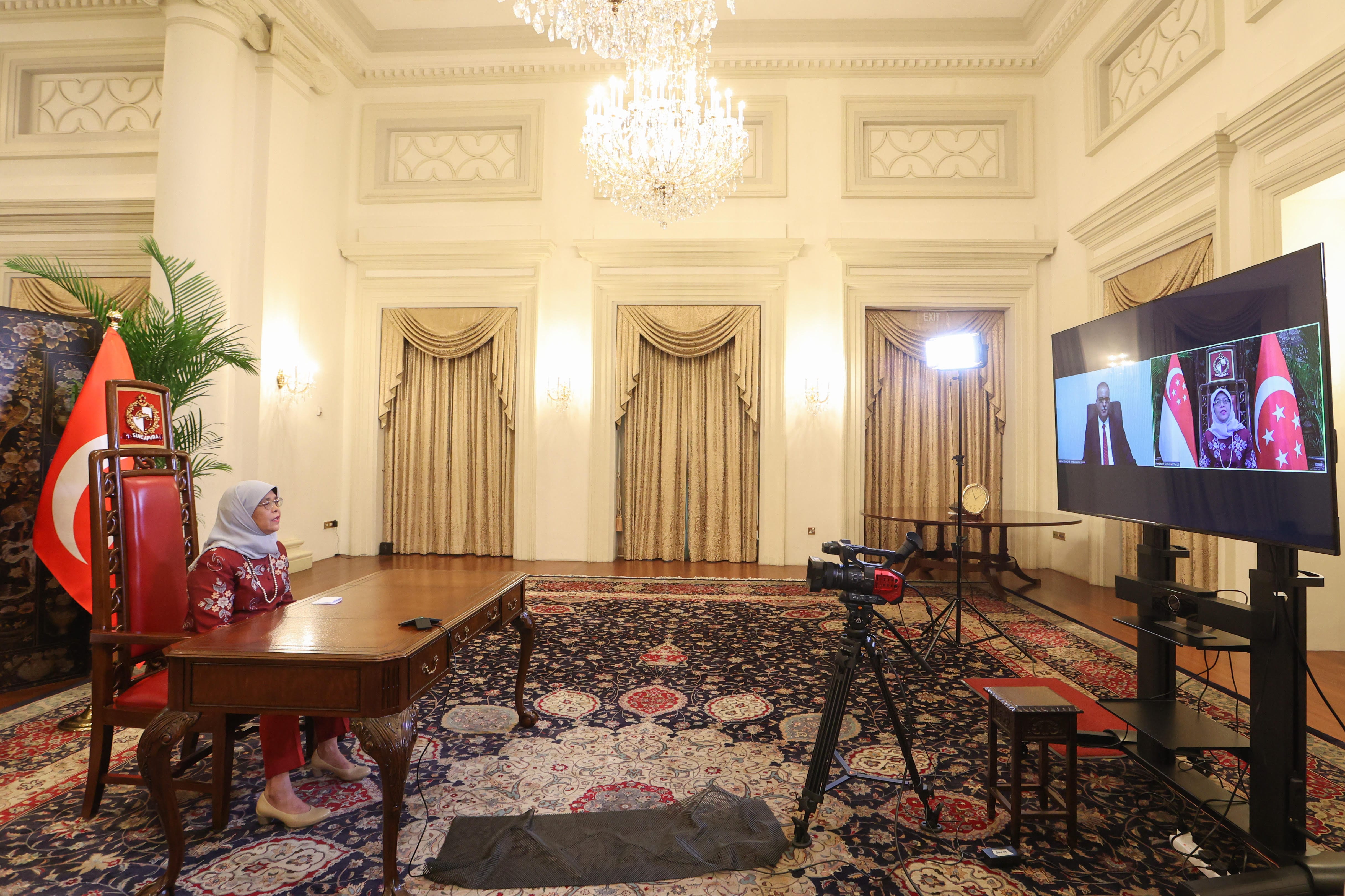 Halimah Yacob on a screen with dignitaries, flags, in an ornate hall with a camera.