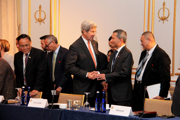 John Kerry shaking hands with a man at a table labeled "Vietnam". Other attendees visible.