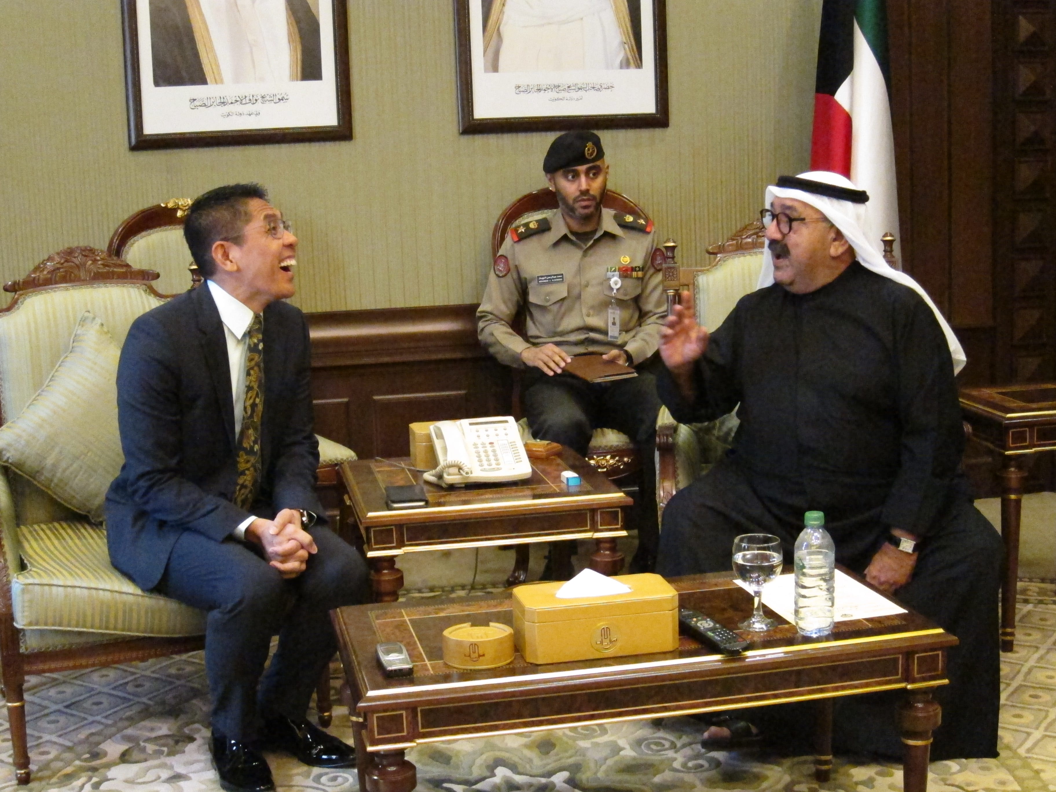 Two men in discussion, one in business suit, one in traditional Kuwaiti attire, plus a guard in uniform.