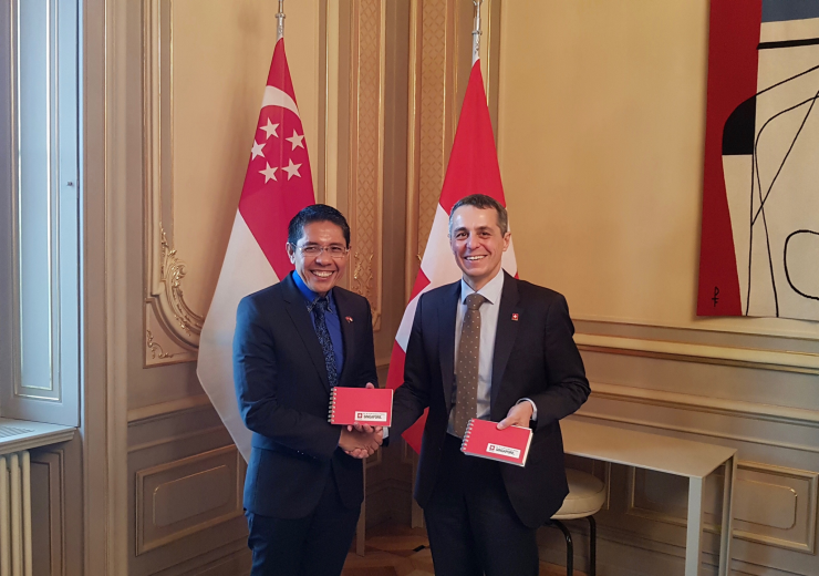 Two men in suits shake hands, holding "Singapore" branded notebooks, with Singapore & Switzerland flags behind.